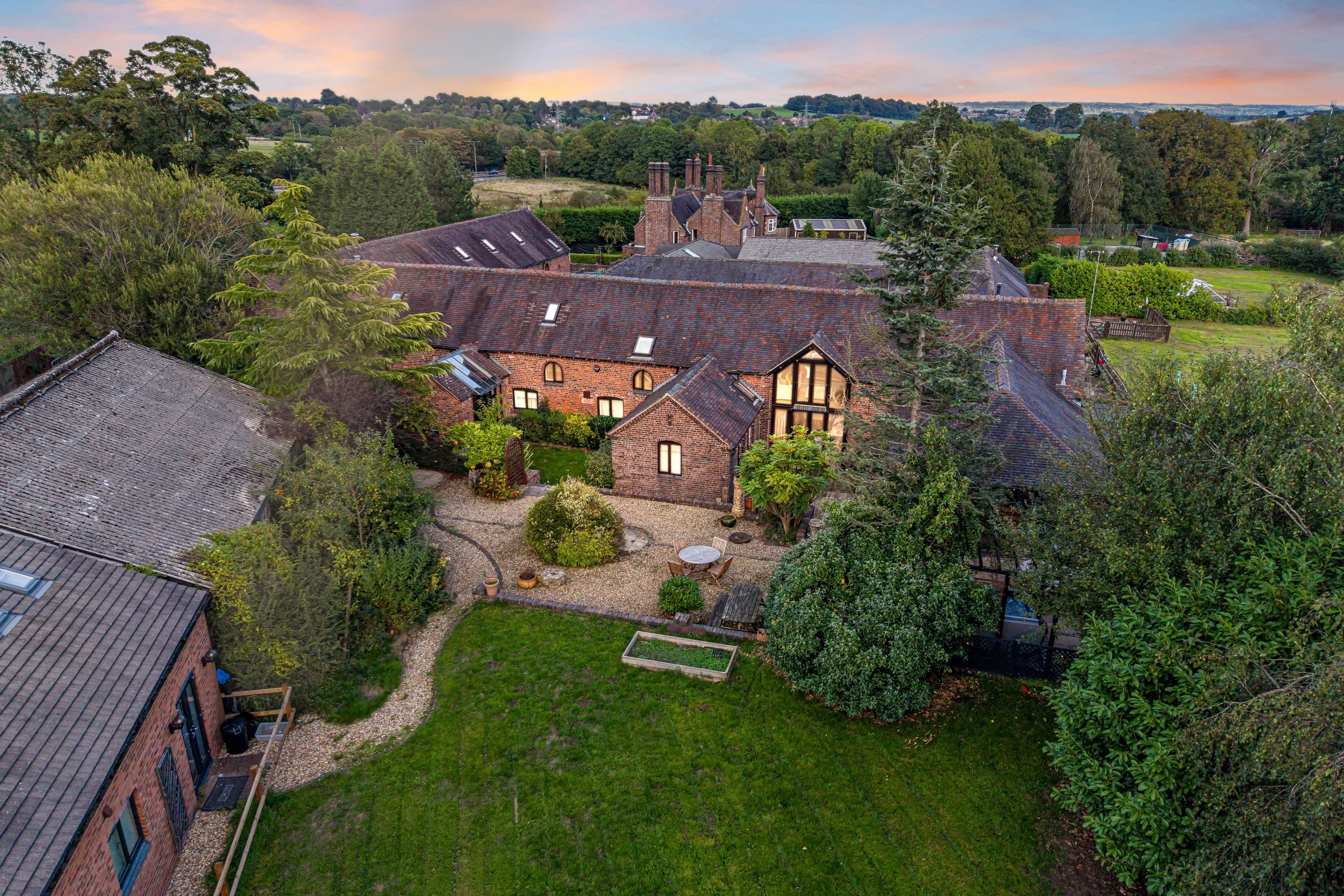 A view of a residential backyard with a brick house, a small patio area with chairs and a circular table, lush green trees and bushes, and a view of a countryside landscape with rolling hills and scattered houses in the distance during sunset.