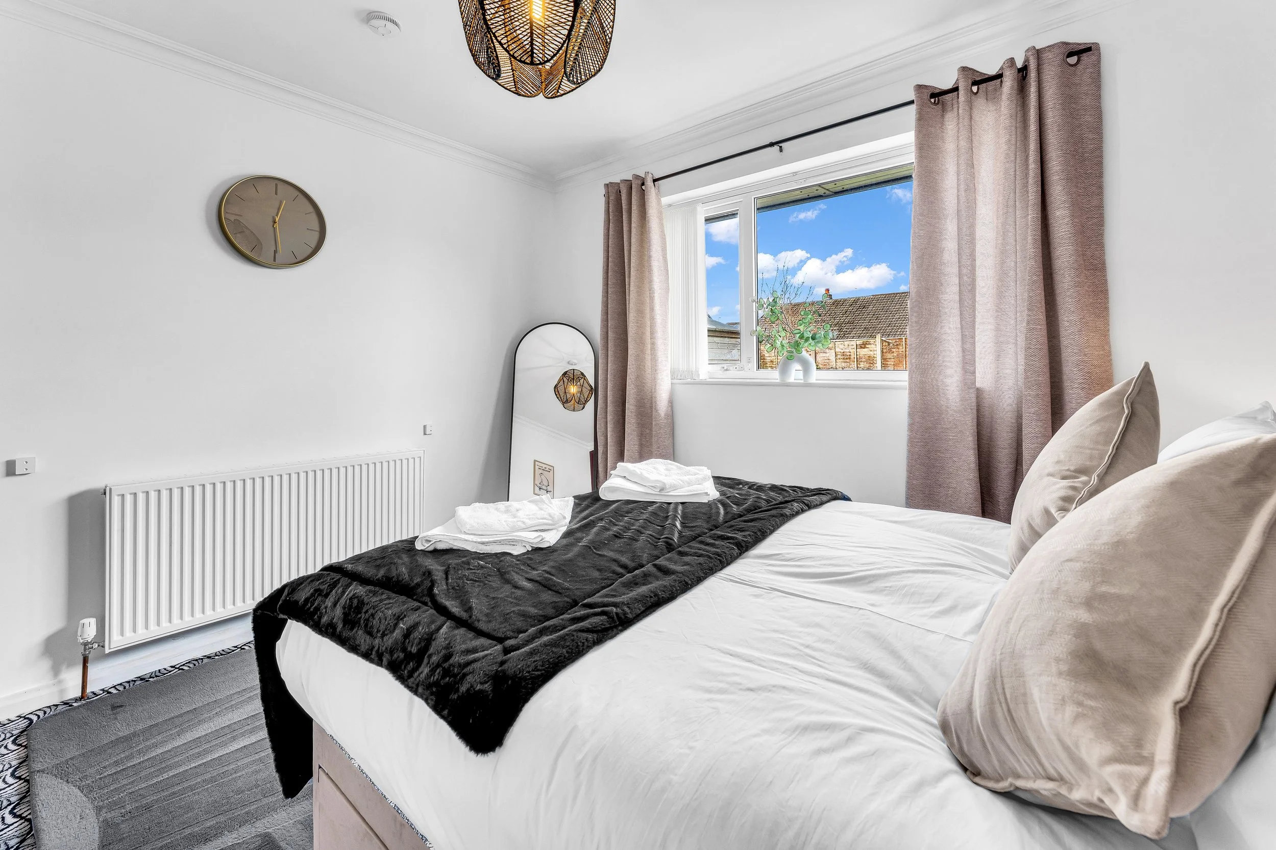 A neatly made bed in a bright bedroom with a window showing a blue sky and neighborhood. The room has beige pillows, a black throw blanket, folded towels, a round mirror, a wall clock, a floor mirror, a vase with greenery, beige curtains, and a decor