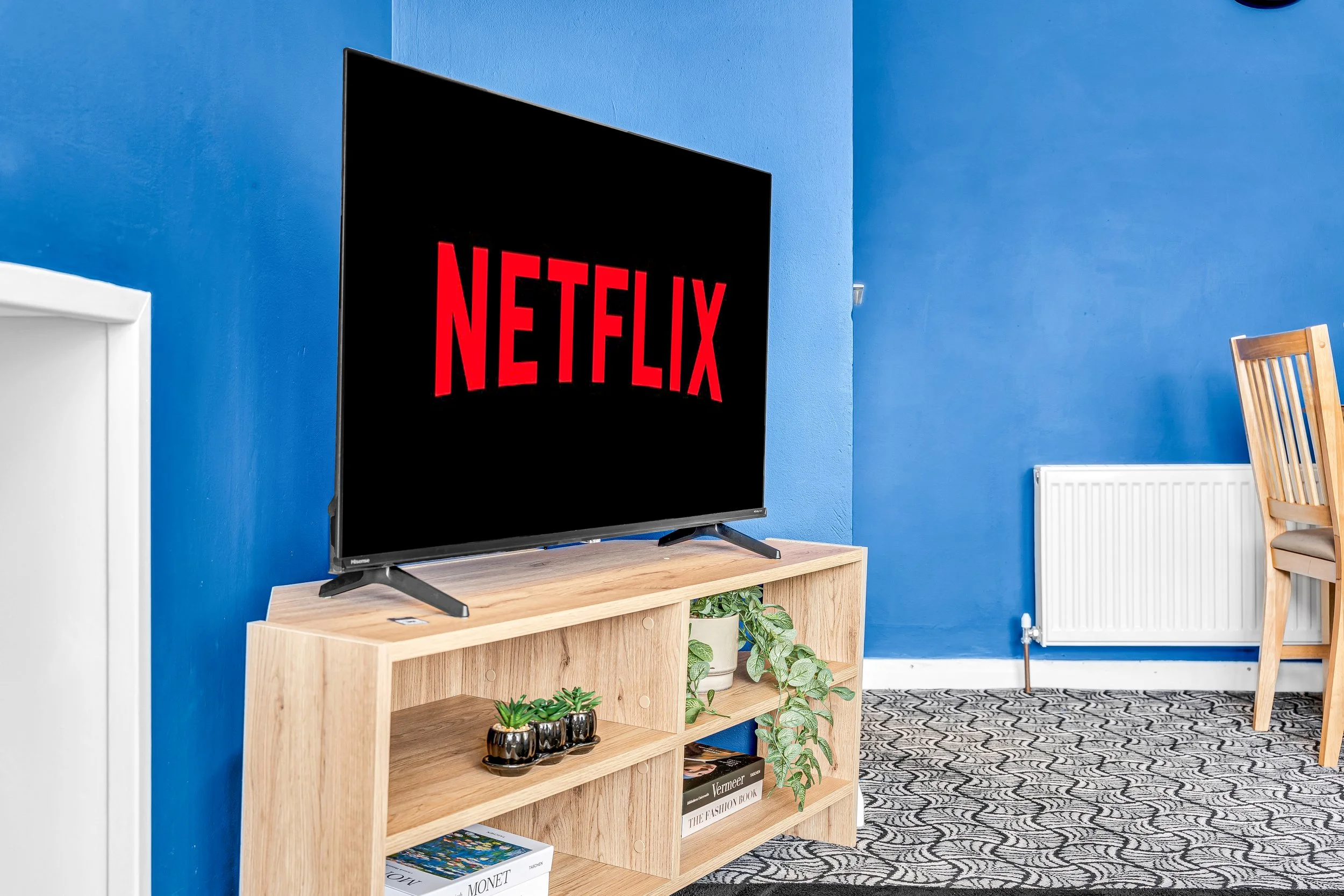 A flat-screen TV displaying the Netflix logo in red text on a black background, placed on a wooden stand with plants and books, in a room with bright blue walls and a patterned carpet.
