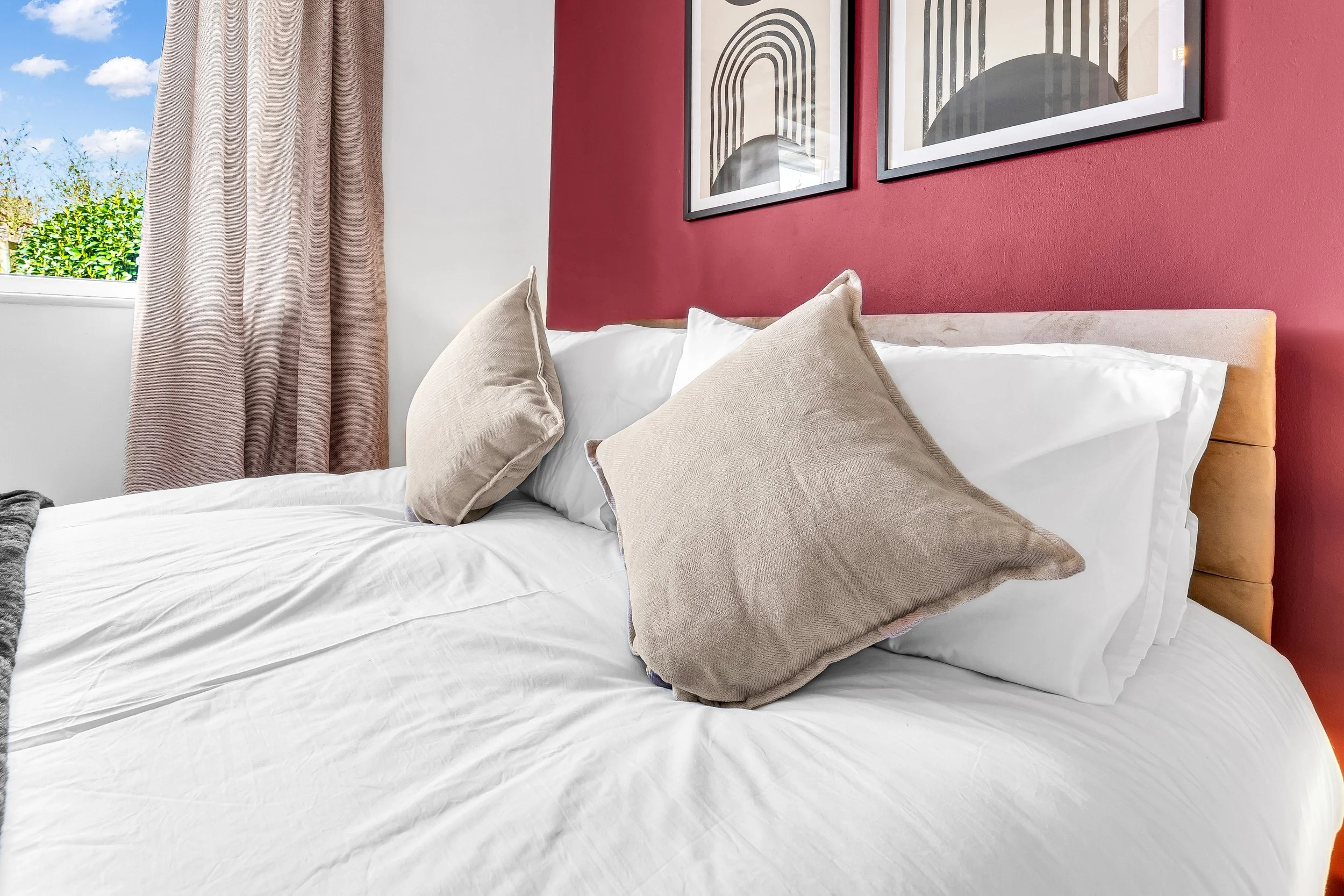 Close-up of a bed with white sheets, beige pillows, and a tan headboard, against a red accent wall with two black-and-white geometric framed artworks. A window with beige curtains and blue sky with clouds is visible on the left.