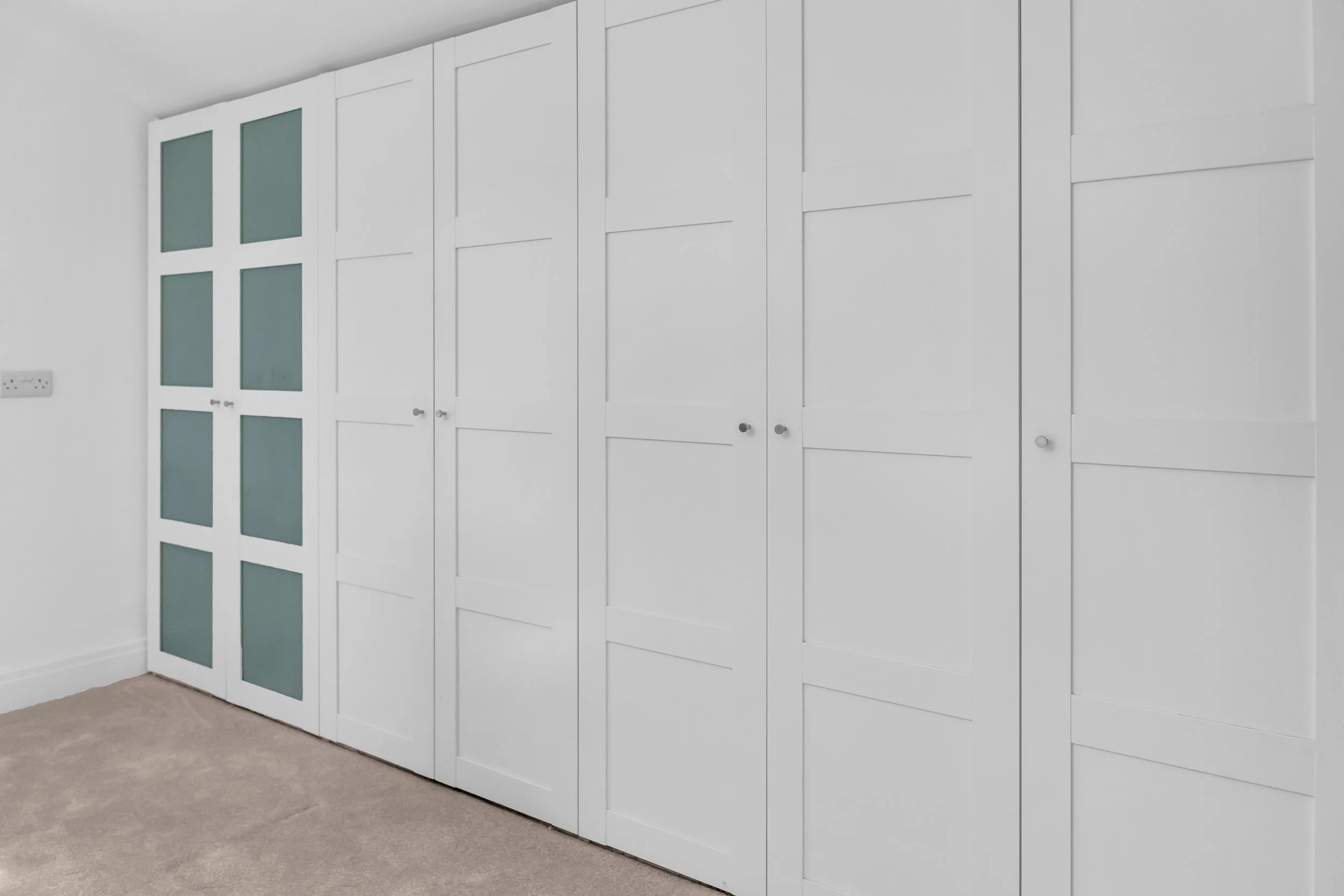 A white wall with multiple built-in white cabinets with doors, some of which have black knobs. On the wall to the left, there are two rectangular glass panels with a frosted appearance. The floor has beige carpet, and there are two electrical outlets