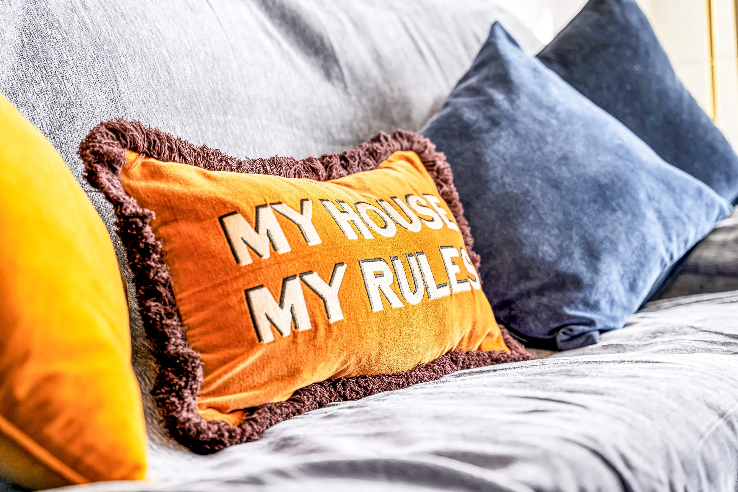 Decorative throw pillows on a sofa, including an orange pillow with dark border that says "My House My Rules" in bold white and black letters, and two blue pillows behind it.