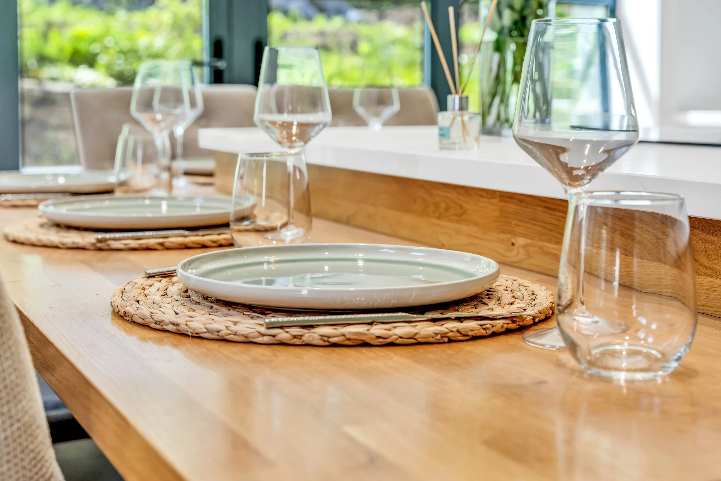 A dining table set with plates, water glasses, and woven placemats, with a window showing greenery outside in the background.