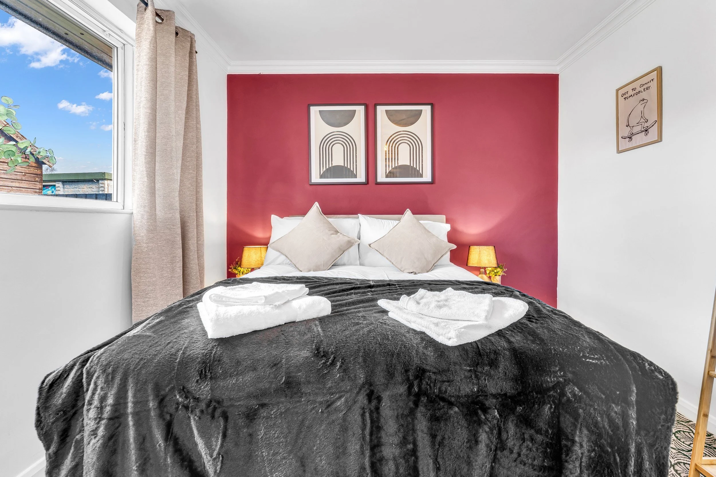 A neatly made bed with white pillows and beige cushions, covered with a black blanket. Towels are folded on the bed. Two small lamps are on bedside tables, placed against a red accent wall with two framed abstract art prints. A window with beige curt