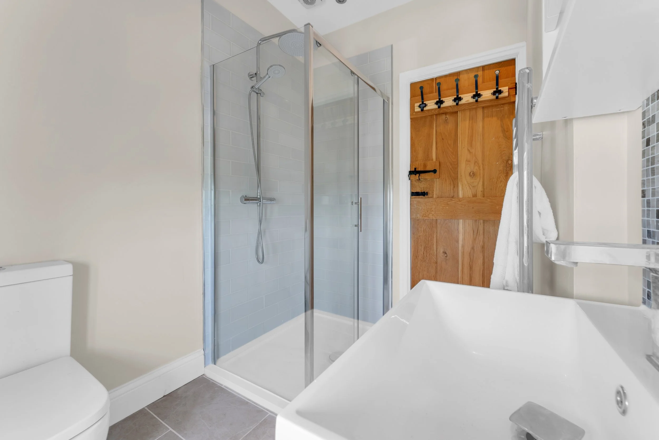 A small bathroom featuring a glass shower enclosure with two doors, a white toilet, a bathtub, a towel rack with a white towel, and a wooden door with black hooks. The walls are painted light, and there are small grey tiles on the floor.