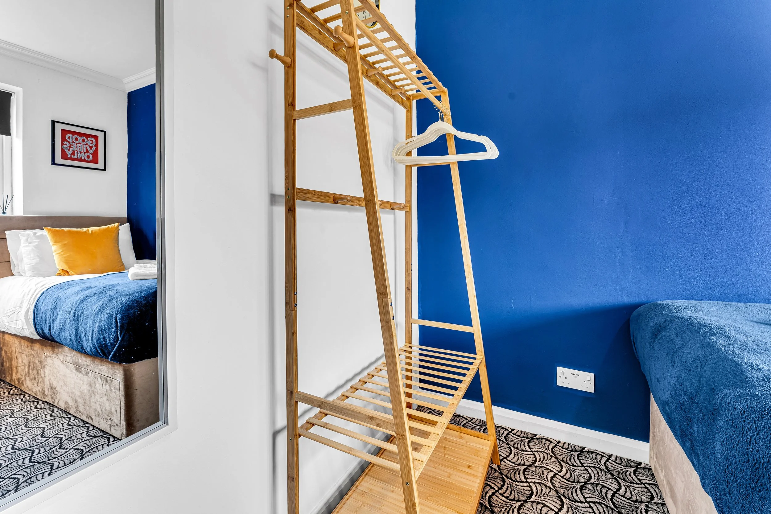 A bedroom with a blue accent wall, a bed with white, blue, and yellow pillows, and a wooden clothing rack with a white hanger.