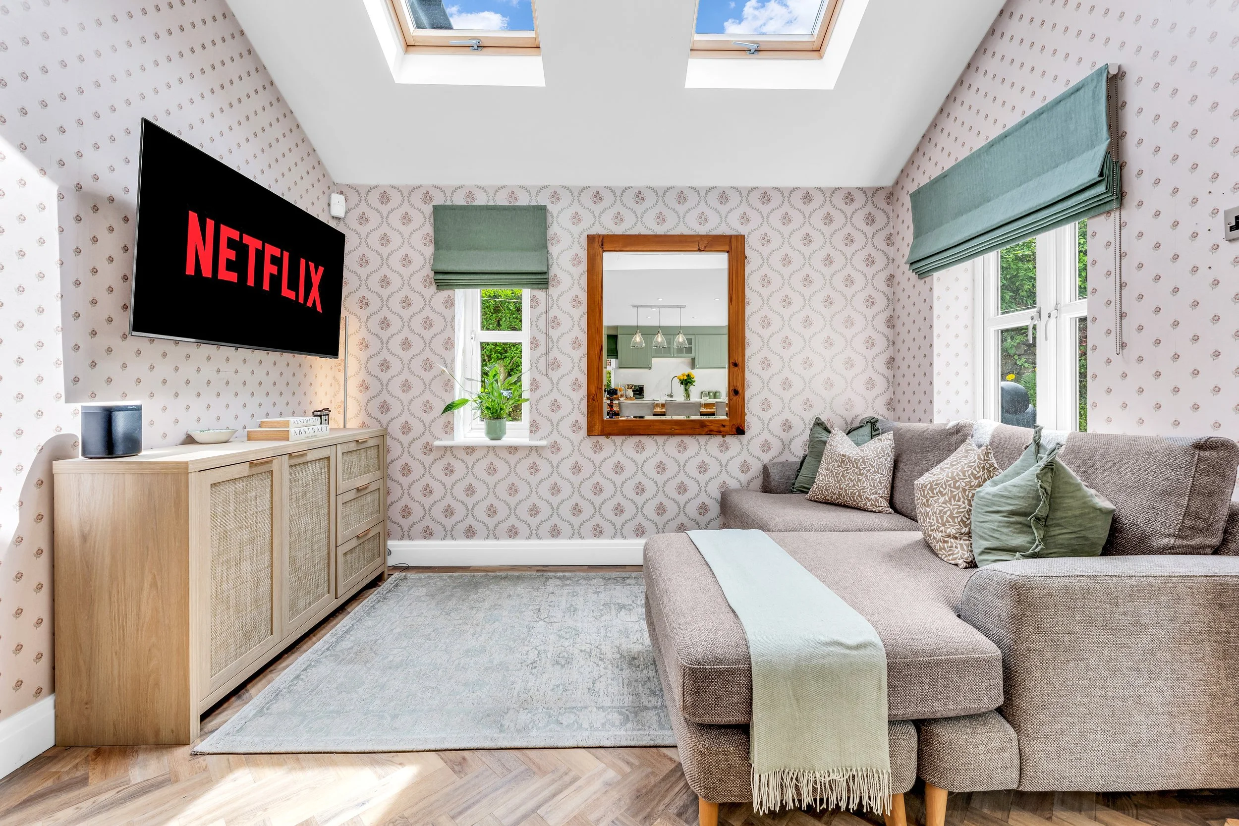 Living room with wallpapered walls, a gray sectional sofa with patterned pillows, a gray rug, a wooden console with a speaker and books, a potted plant, a mirror, and windows with green Roman shades. Skylights show a blue sky with clouds.