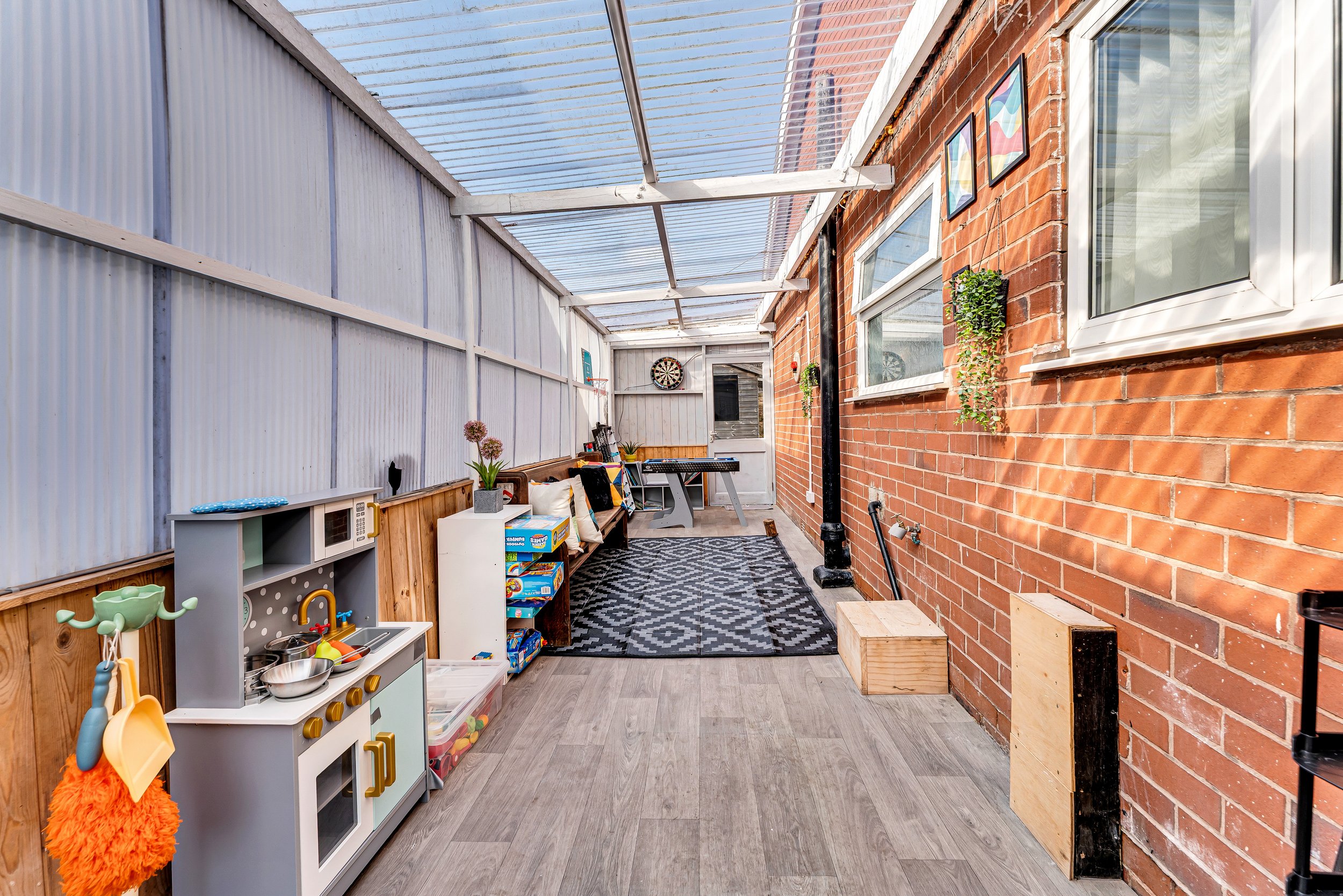 Enclosed outdoor patio with a children’s play kitchen, books, and a dartboard, featuring brick and wood walls, framed art, and a black and white patterned rug.