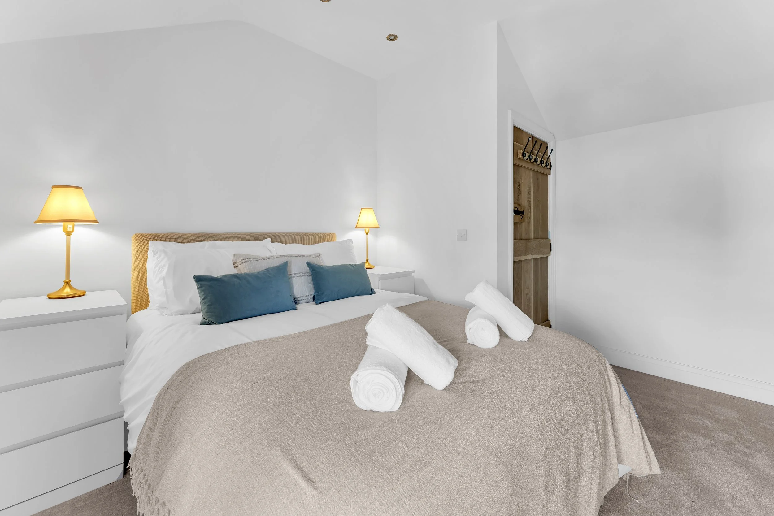 A neatly made bed with white and beige bedding, decorated with blue pillows, and topped with rolled white towels in a minimalistic bedroom. Two yellow lamps provide soft lighting, and there is a small white nightstand on one side. A wooden door with 