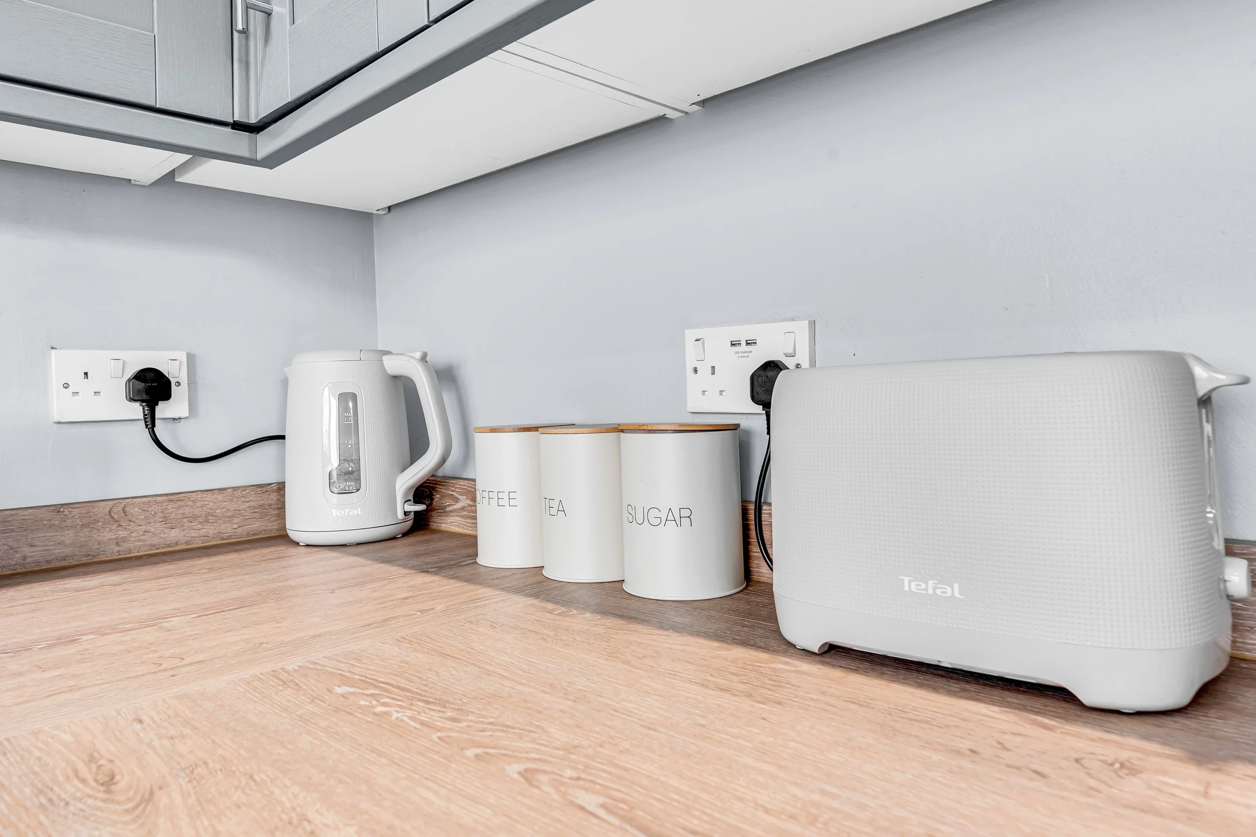 Kitchen countertop with a white electric kettle, four white labeled canisters for coffee, tea, sugar, a white Tefal toaster, and two electrical outlets.