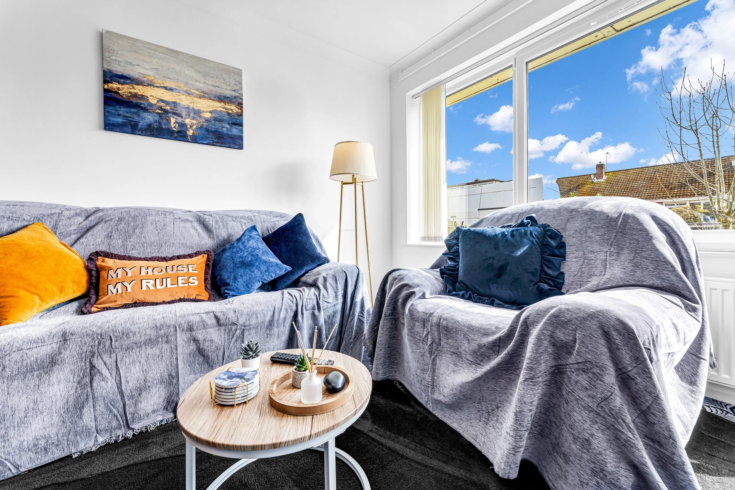 Living room with gray sofas, colorful pillows, a small round wooden table with decor, large window with a tree outside and clear blue sky, white walls with a painting.