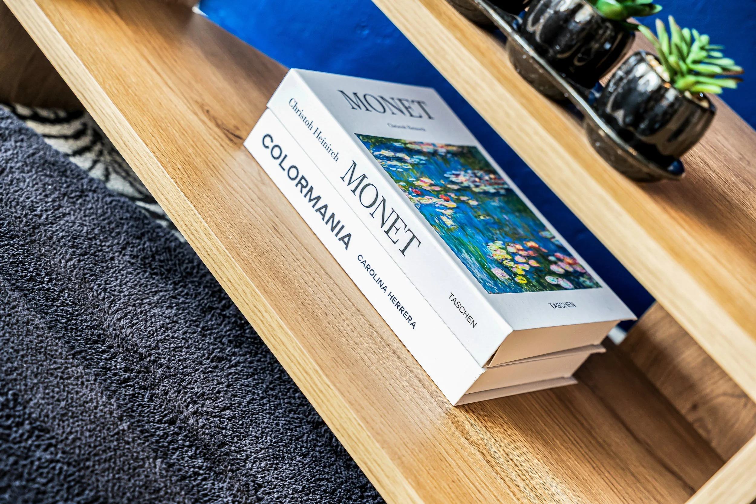 Stack of art books on a wooden table, including a book about Monet and a book about Colormania, with a glass vase containing small plants nearby.