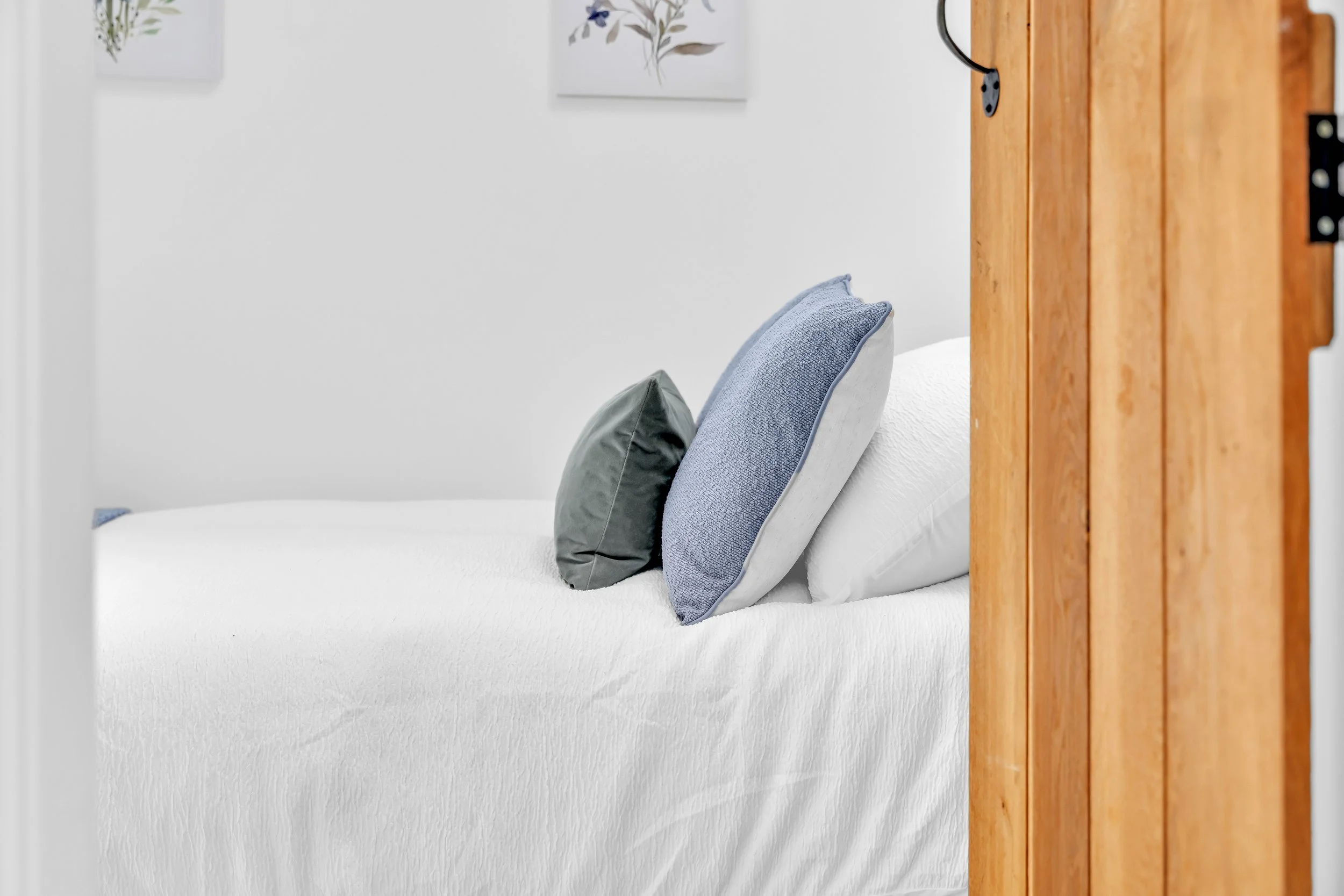 A neatly made bed with three pillows in varying shades of gray and blue, against a white wall with two framed botanical art prints. A wooden door or panel partially visible on the right side.