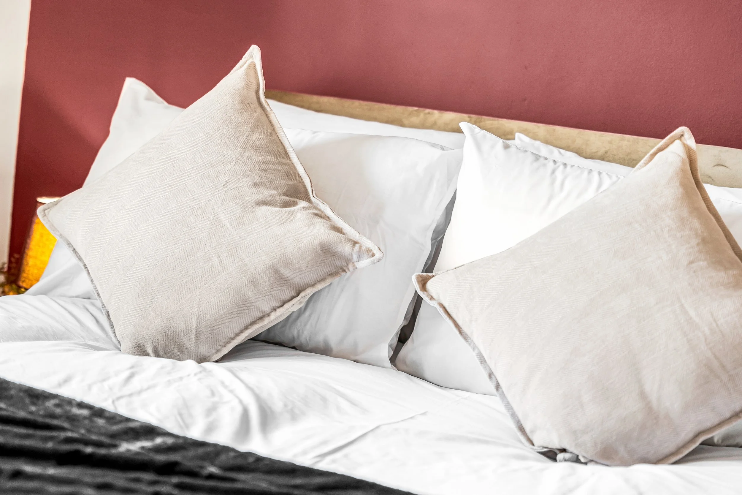 Made bed with white sheets, beige pillows, beige throw pillows, wooden headboard, and red wall.