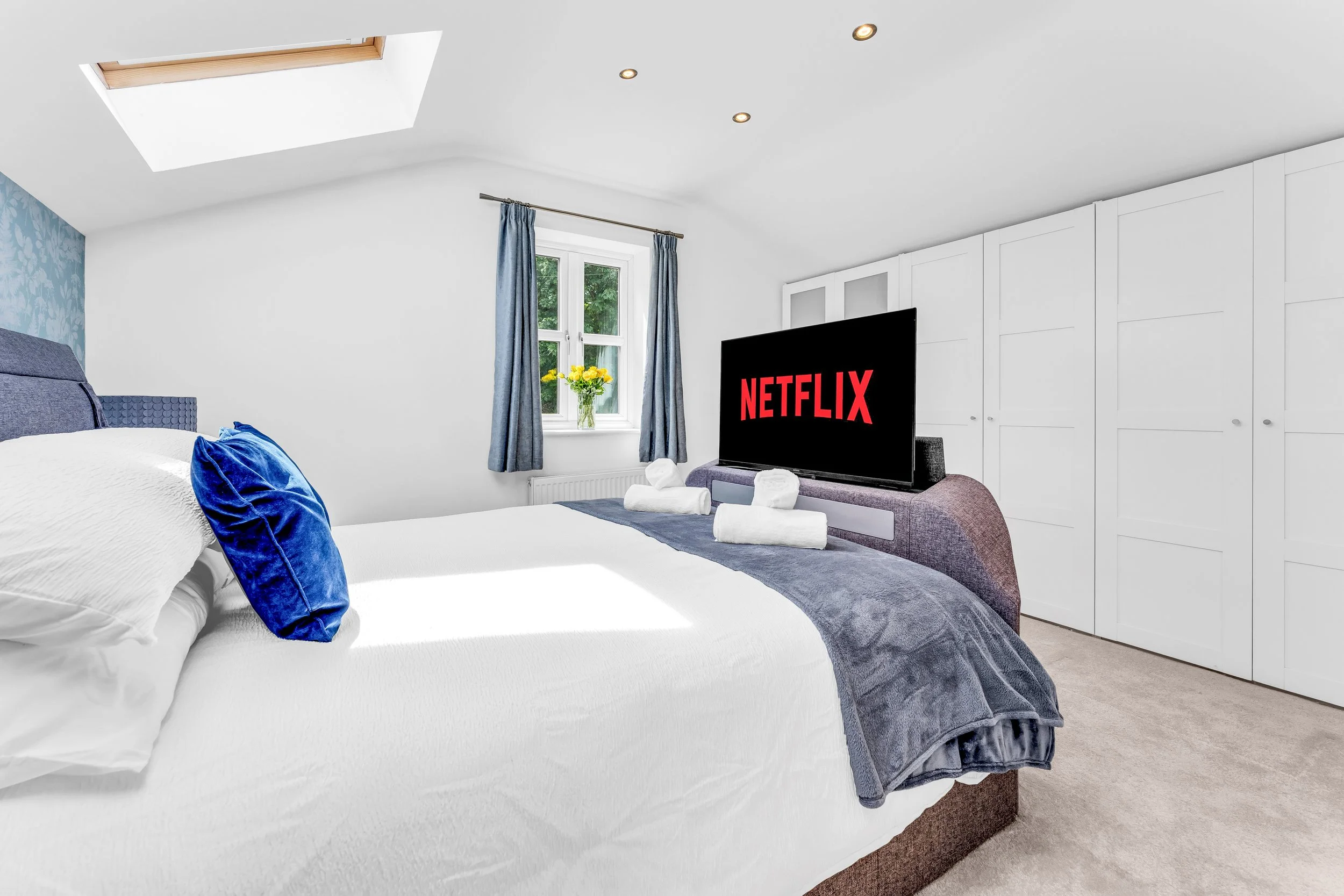 A bedroom with a white bed, blue pillows, a window with blue curtains, a TV displaying the Netflix logo, a vase of yellow flowers, and a closet with white doors.