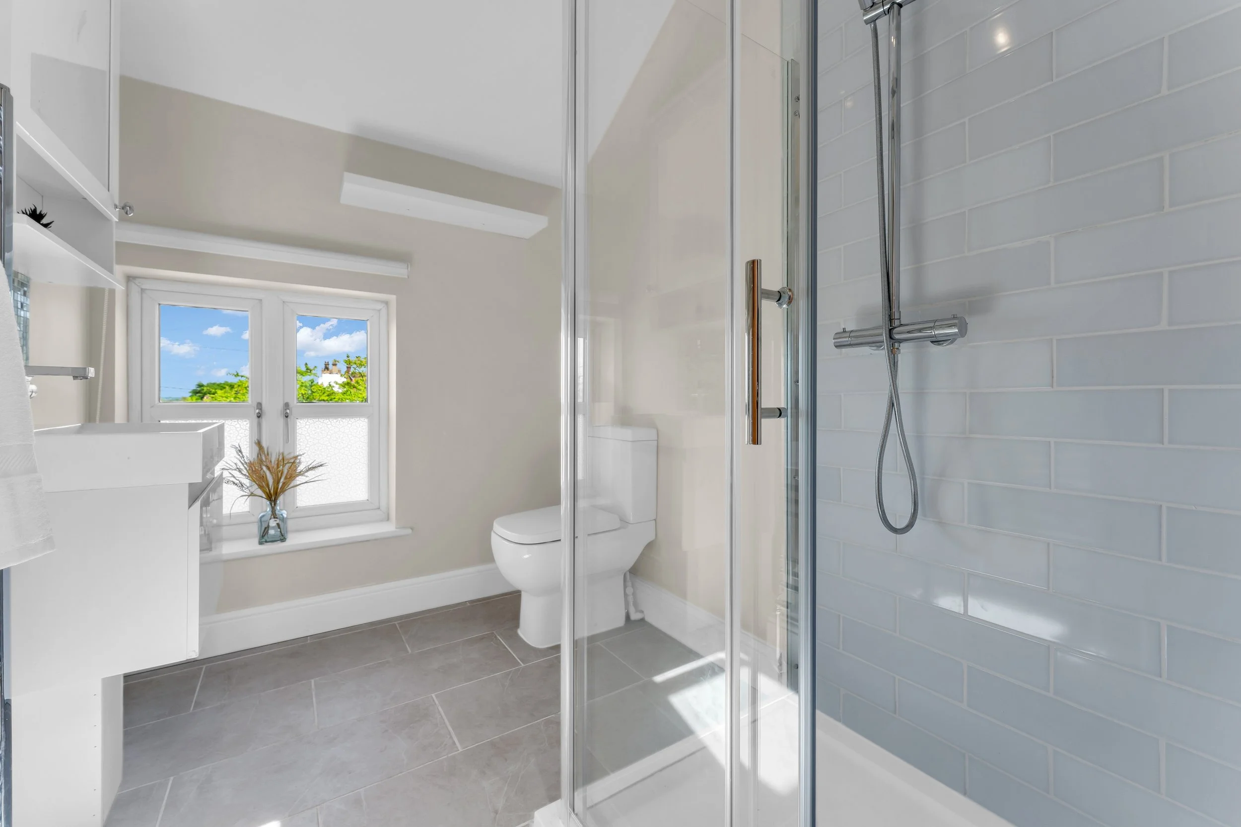 Bright bathroom featuring a glass shower enclosure, a white toilet, and a window with a view of green trees and a blue sky.