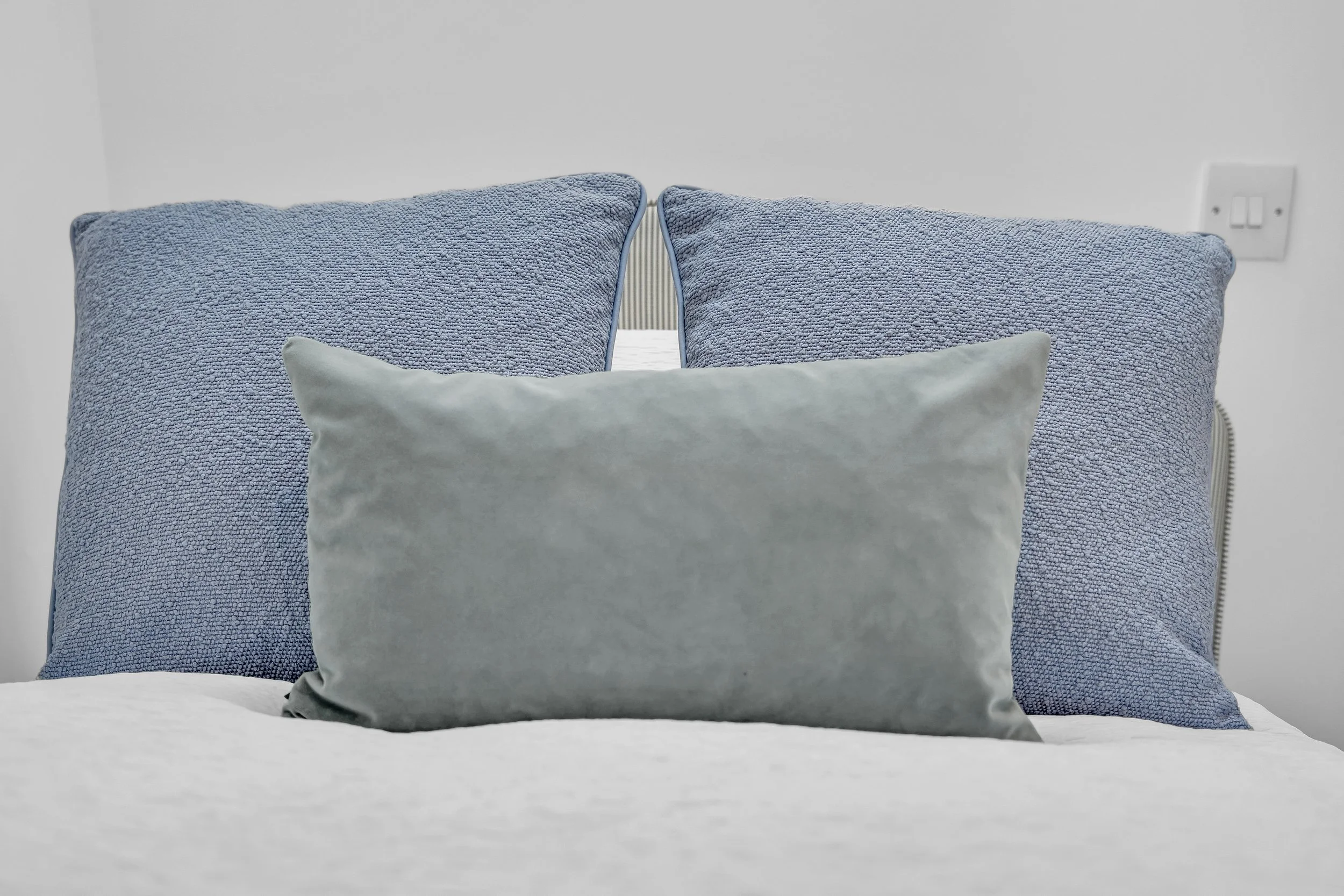 Close-up of a bed with two large light blue pillows and one smaller gray pillow in front, against a white wall with electrical outlets visible.