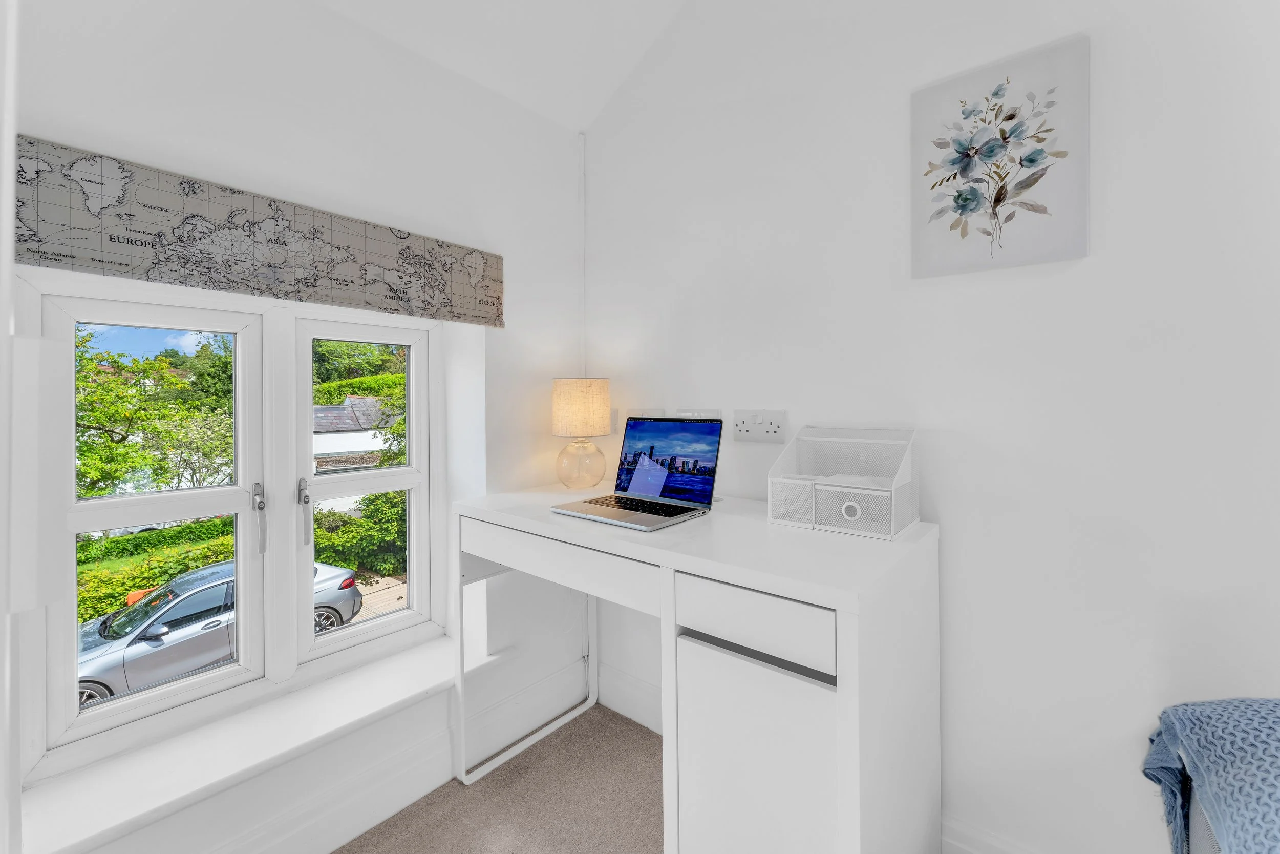A white desk with a laptop, a white lamp, a small notebook holder, and a fluffy blue blanket. Behind the desk is a window showing green trees and a parked gray car outside. The room has white walls adorned with a world map and a floral painting.