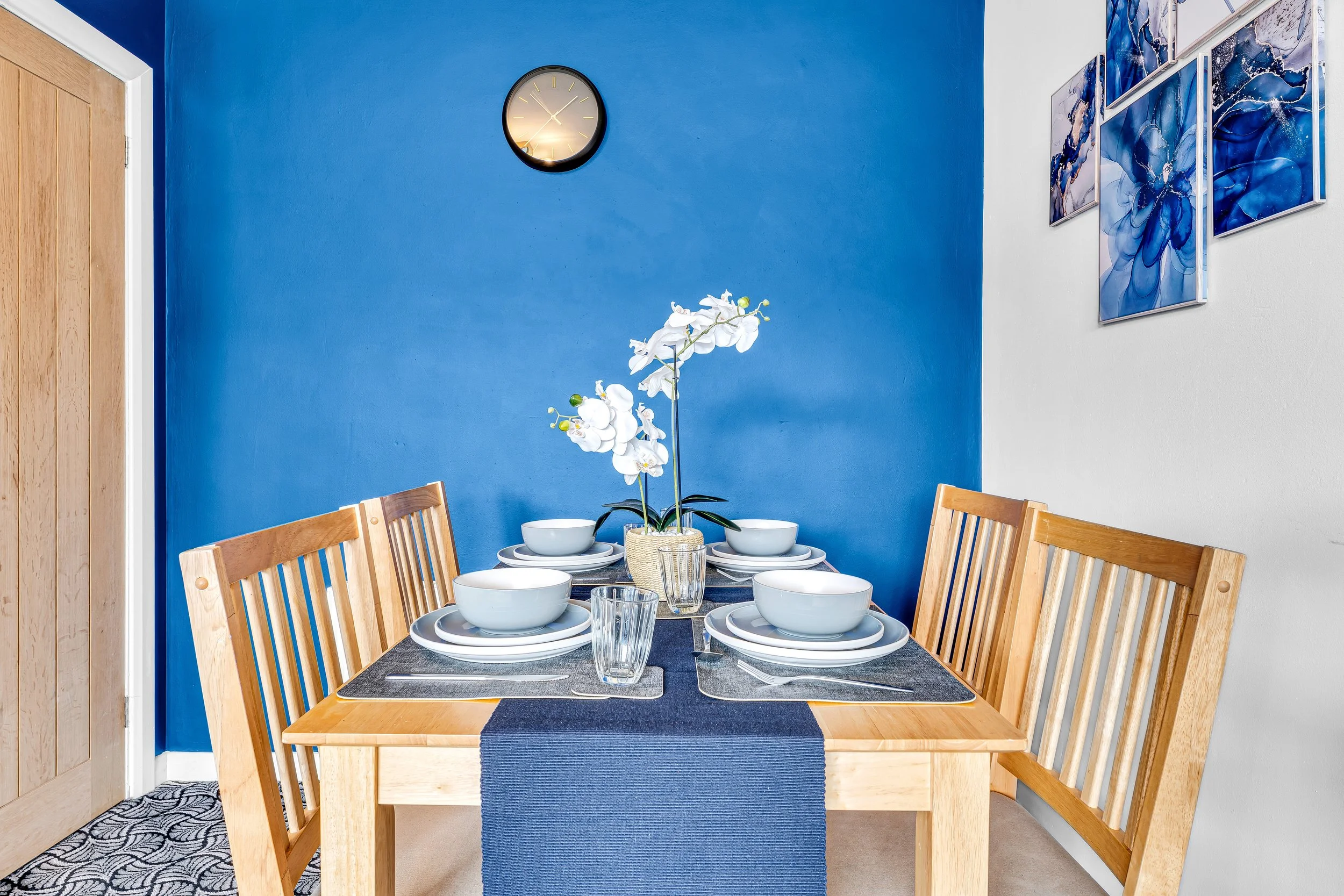 Dining table set for four with white bowls, glasses, and a flower centerpiece against a blue and white wall with artworks and a clock.