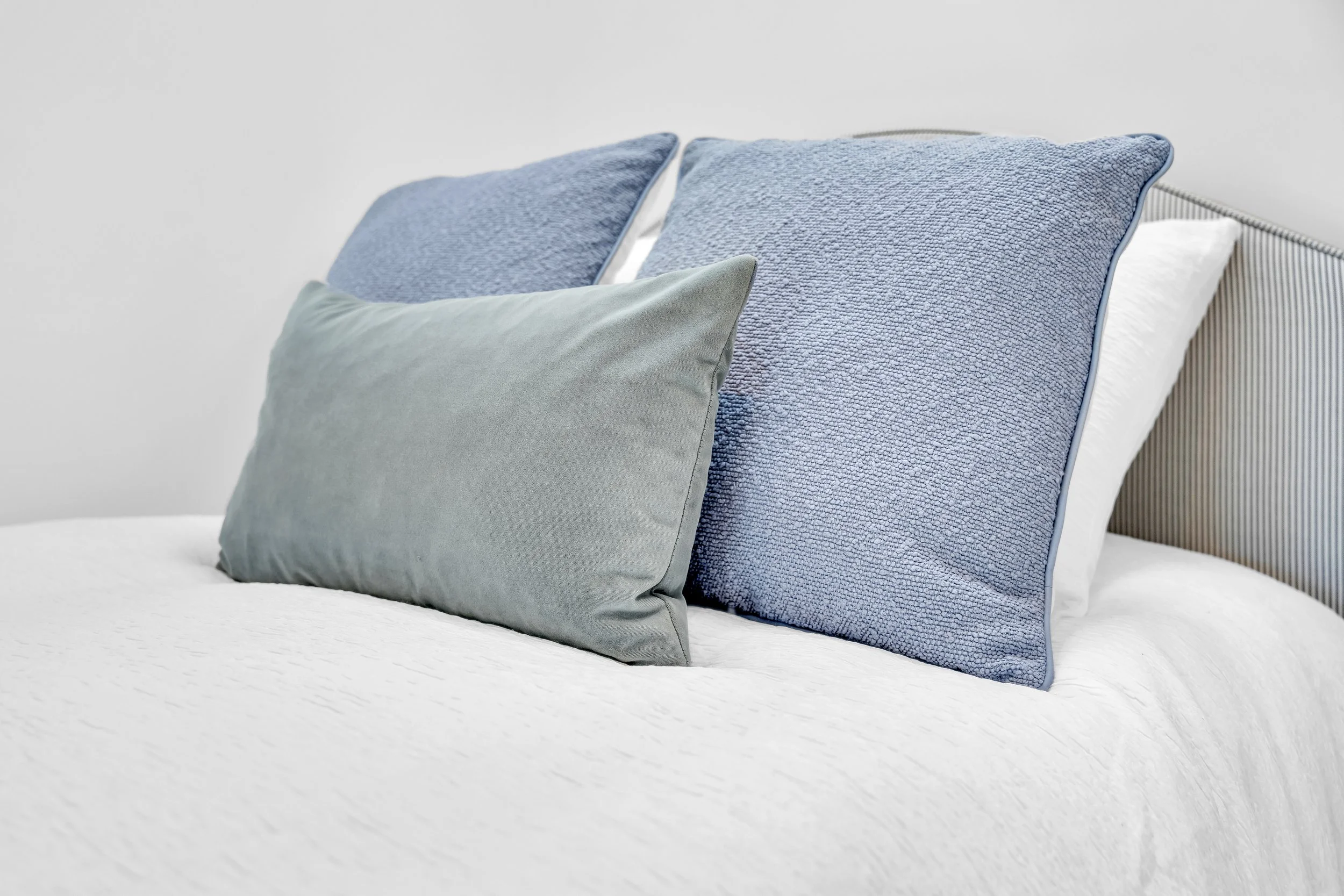 A bed with white sheets and four pillows, two with textured blue covers and one with a plain light gray cover, against a plain white wall.