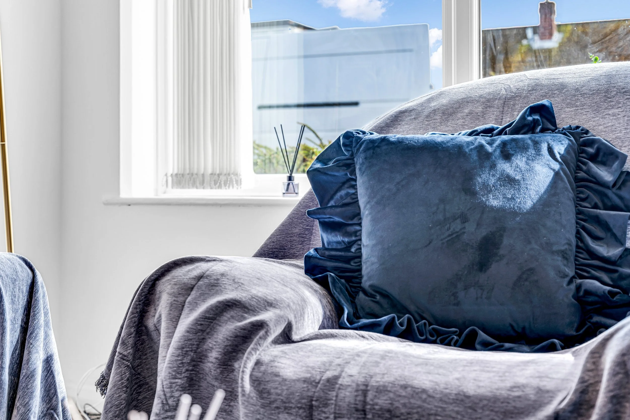 A cozy gray armchair with a plush blue velvet pillow, positioned in front of a window with sheer white curtains, with a vase and reed diffuser on the windowsill outside visible.