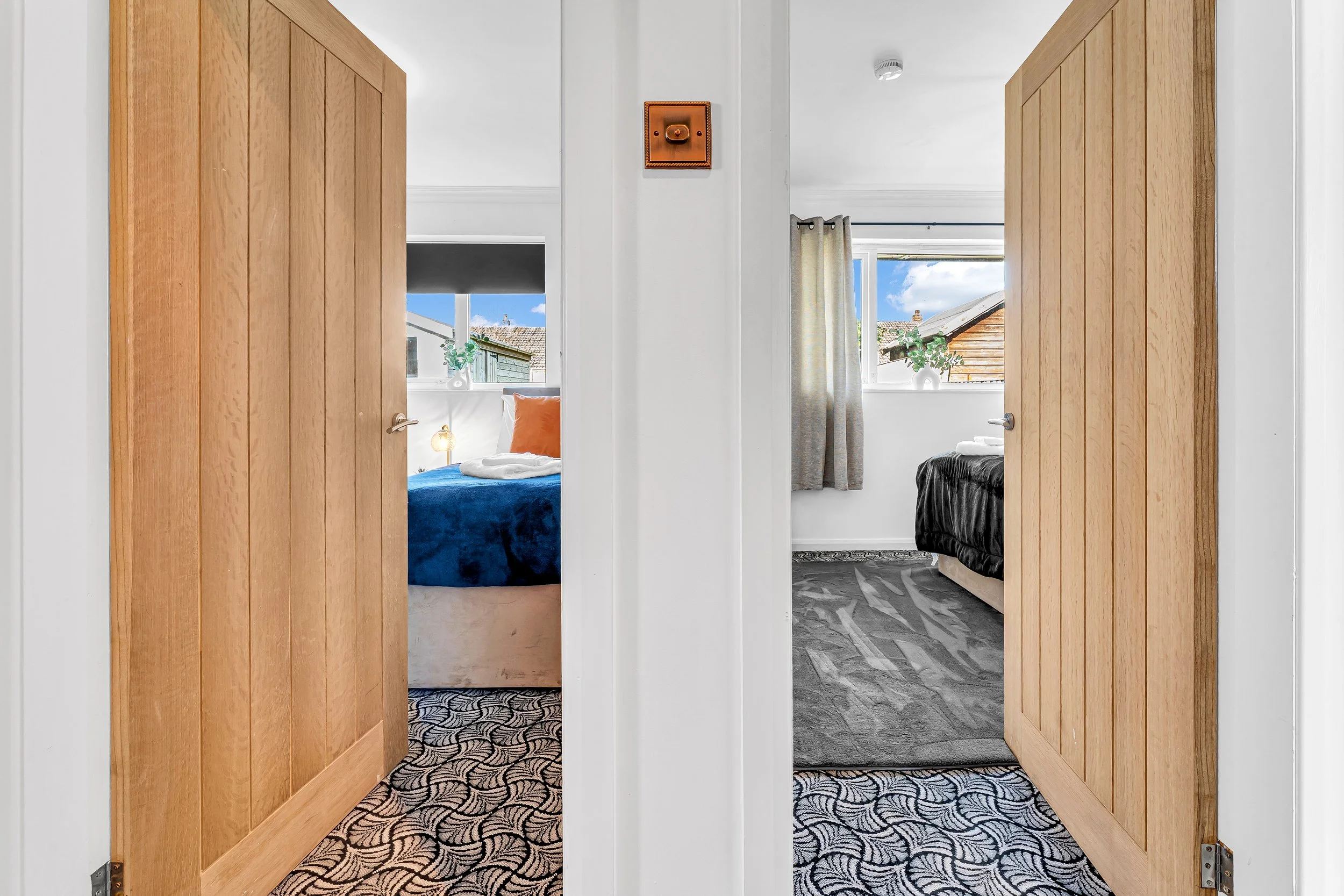 Two open doors reveal separate bedrooms with beds, pillows, and windows showing outside views. One room has a blue bed with a beige base and an orange pillow, and the other has a black bed with white linens. Both rooms have patterned carpets and simp