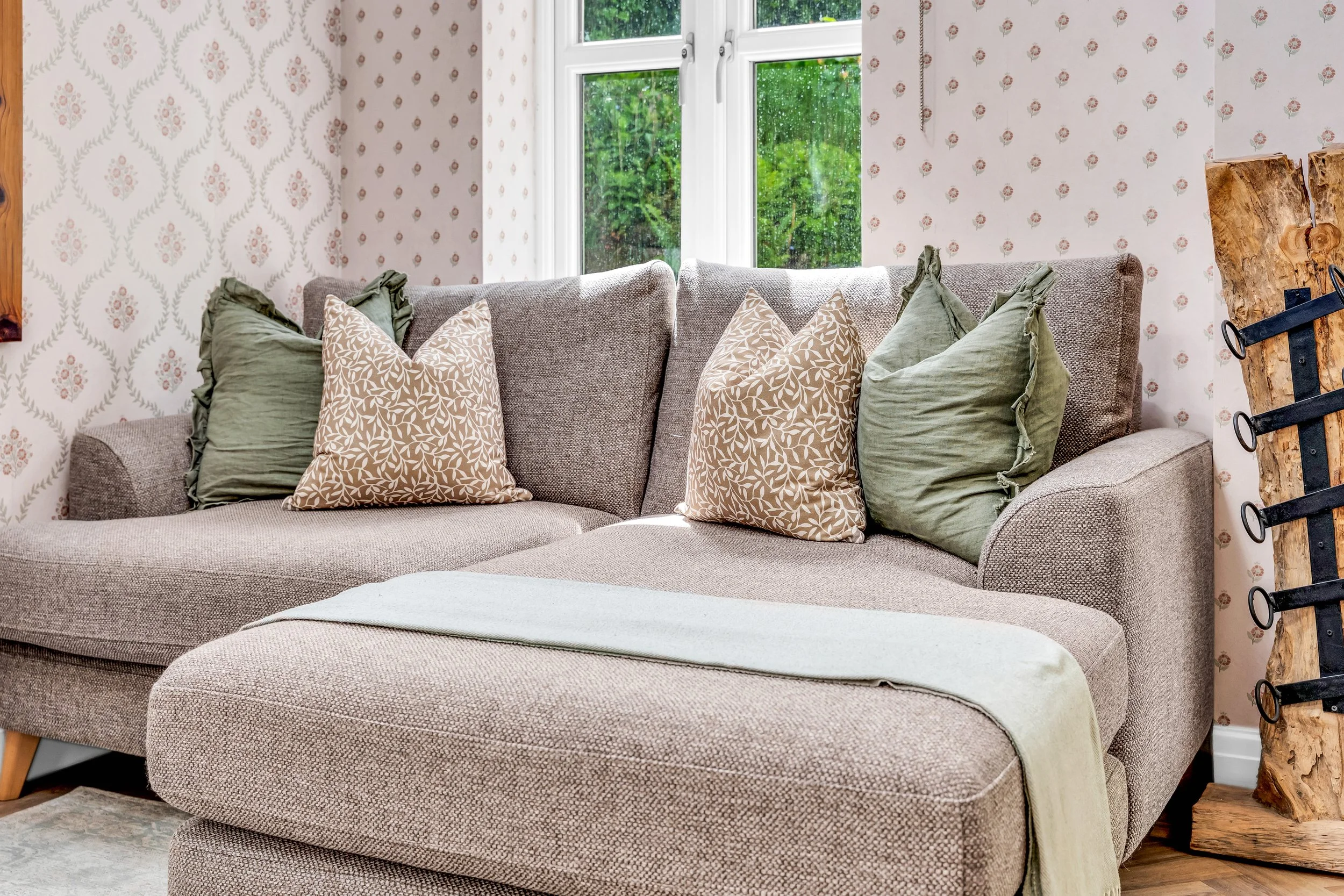 Living room with a beige upholstered sectional sofa and a variety of green and beige patterned pillows, a large window with greenery outside, floral patterned wallpaper, and a rustic wooden log side table.