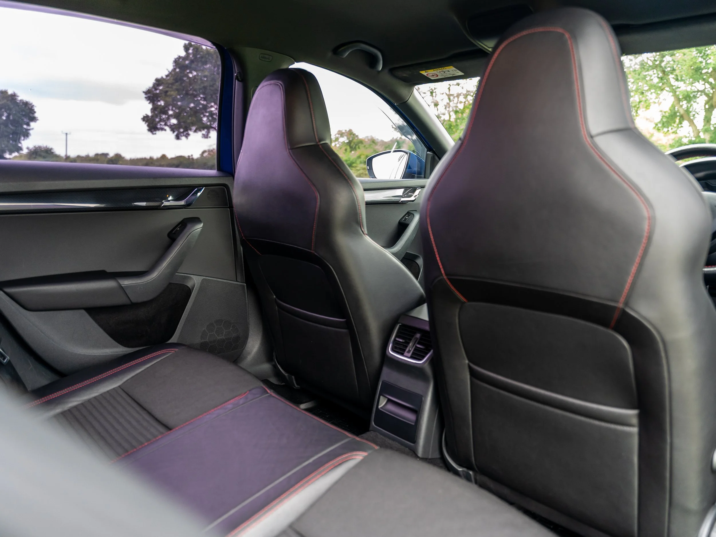 Interior of a modern vehicle showing the back seats and front seats with black leather upholstery and red stitching. The view includes the rear passenger side and the back of the front seats, with an outside landscape visible through the windows.