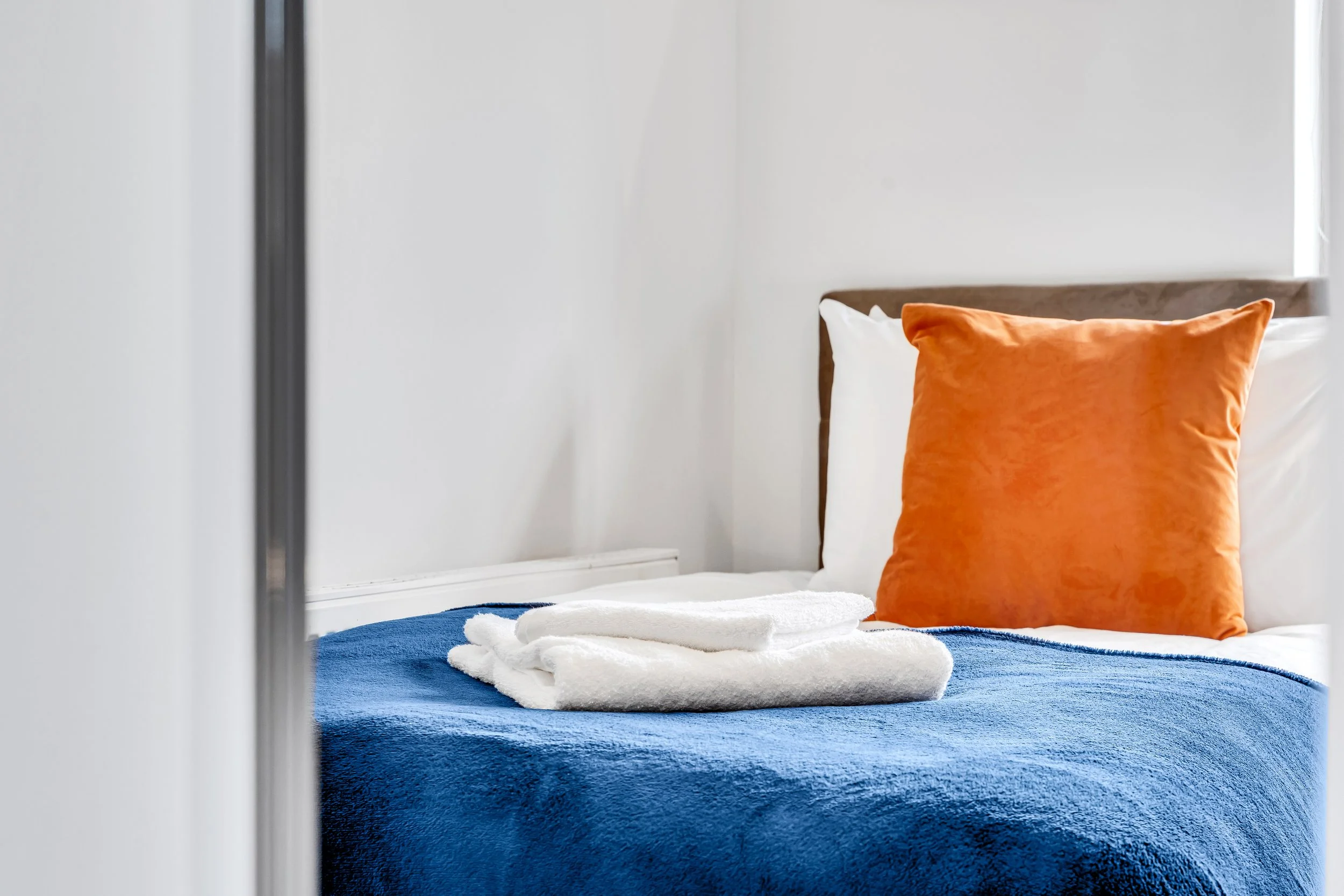 A neatly made bed with a white headboard, blue blanket, white pillows, an orange throw pillow, and white rolled towels on top, in a bright room with white walls.