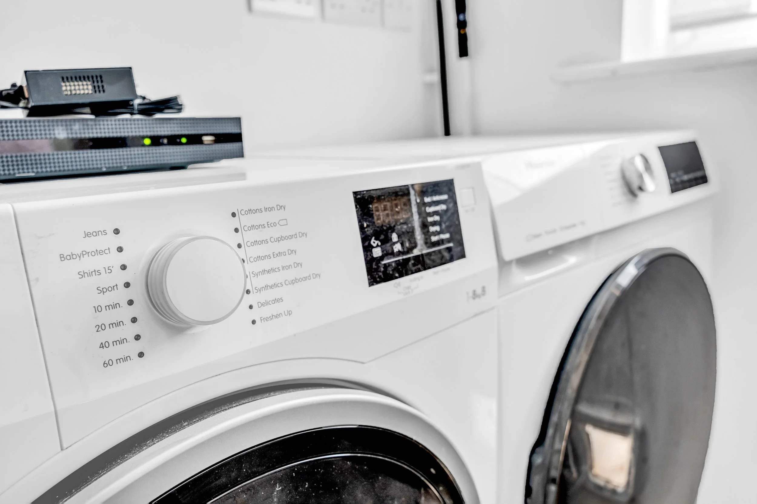 Close-up of a white washing machine with a digital display and control knob, next to another washing machine in a laundry room.