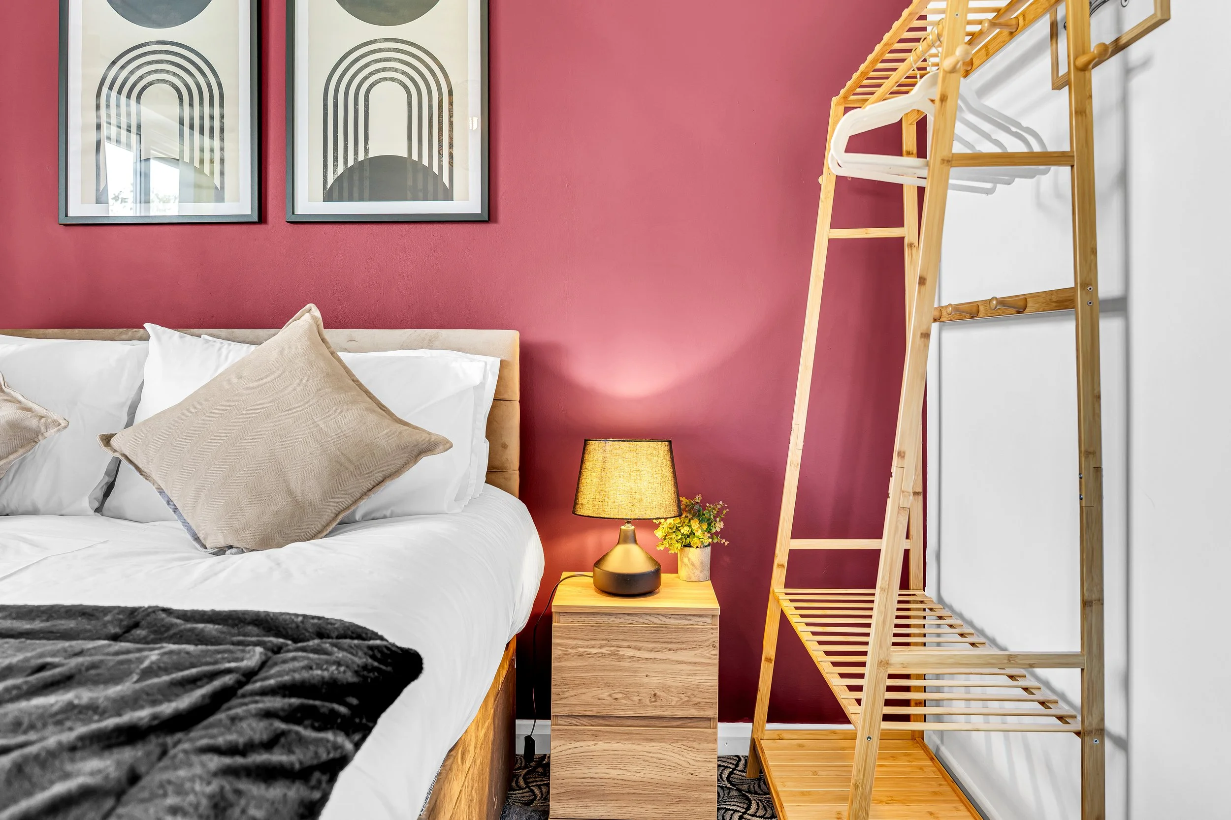 A bedroom with a red accent wall, a bed with white sheets and beige pillows, a nightstand with a lamp and a small potted plant, and a wooden clothing rack.