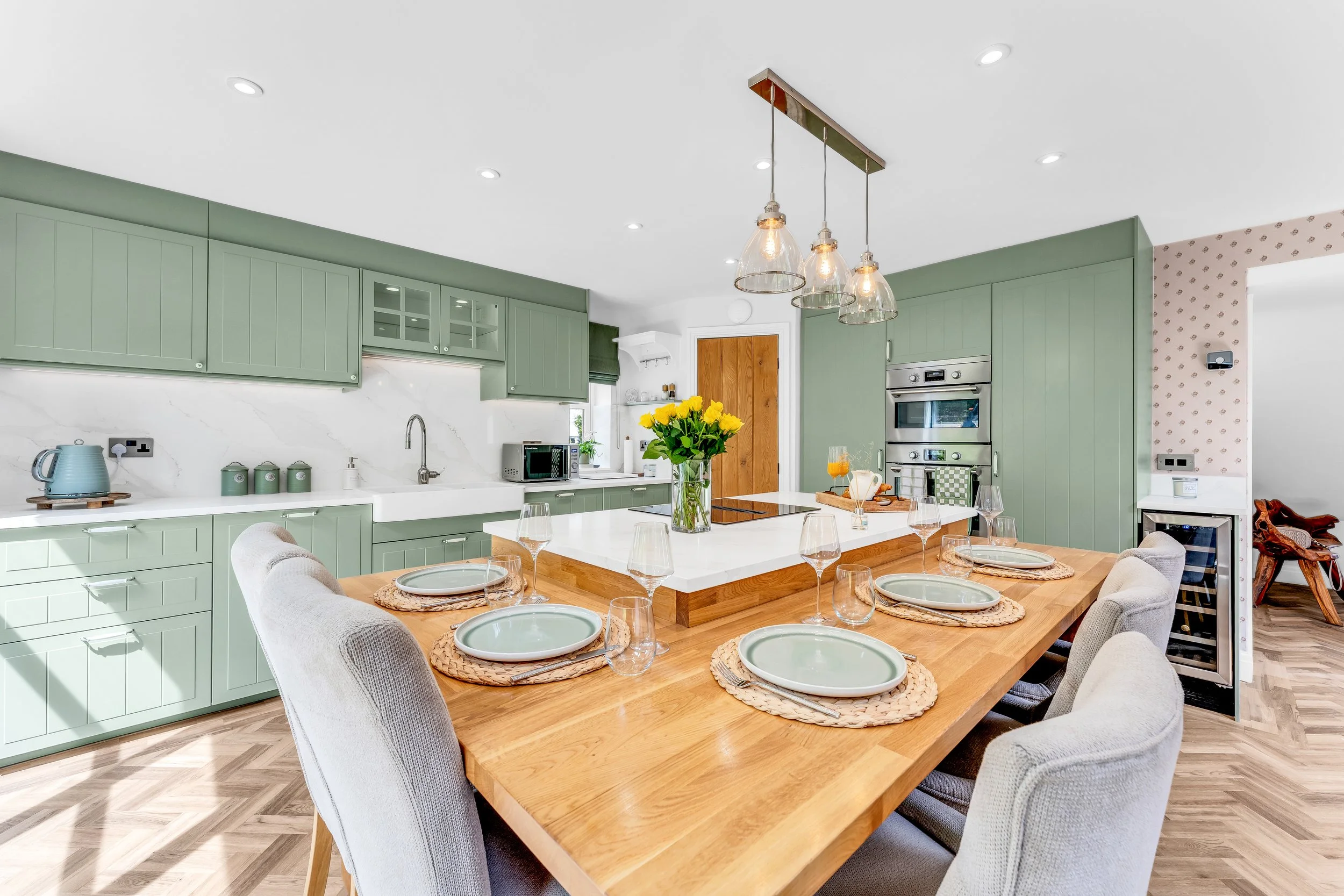A bright, modern kitchen and dining area with sage green cabinetry, a white marble backsplash, and a wooden dining table set with placemats, plates, glasses, and a vase of yellow flowers. The space features pendant lights, stainless steel appliances,