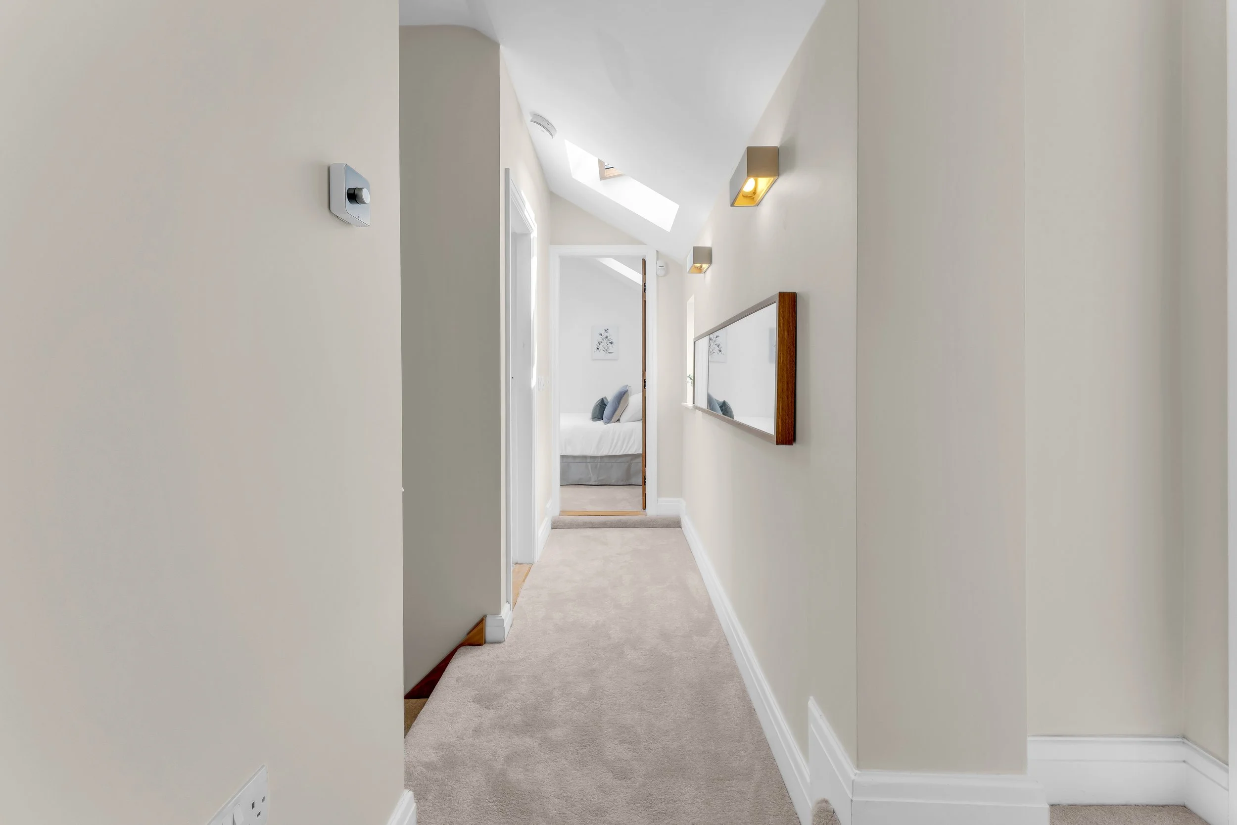 A hallway with beige walls, ceiling lights, and a carpeted floor, leading to a bedroom with a bed, pillows, and artwork on the wall.