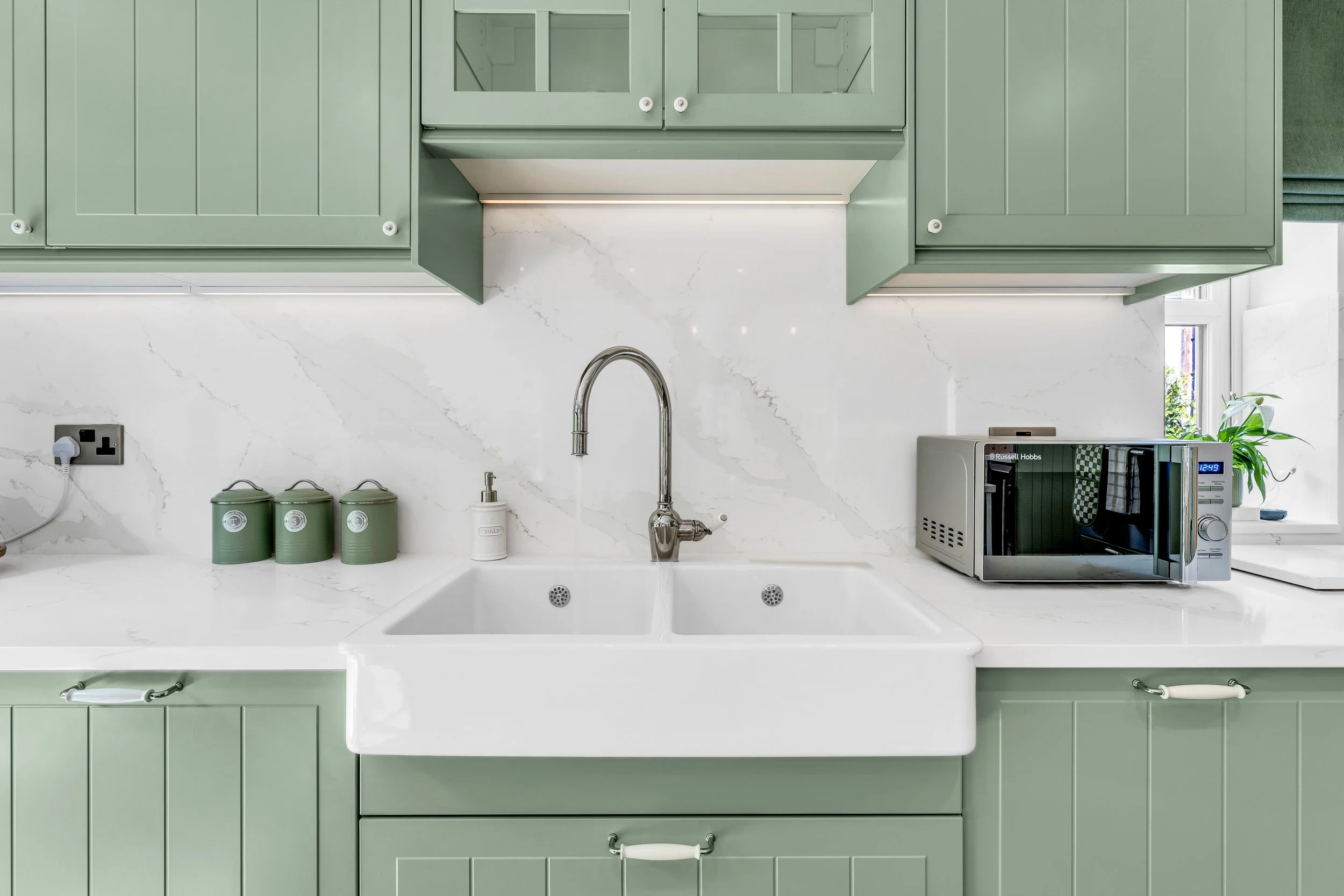 Modern kitchen with green cabinets, white marble countertop, farmhouse sink, stainless steel faucet, three green storage canisters, soap dispenser, microwave oven, and a window with a view of greenery.