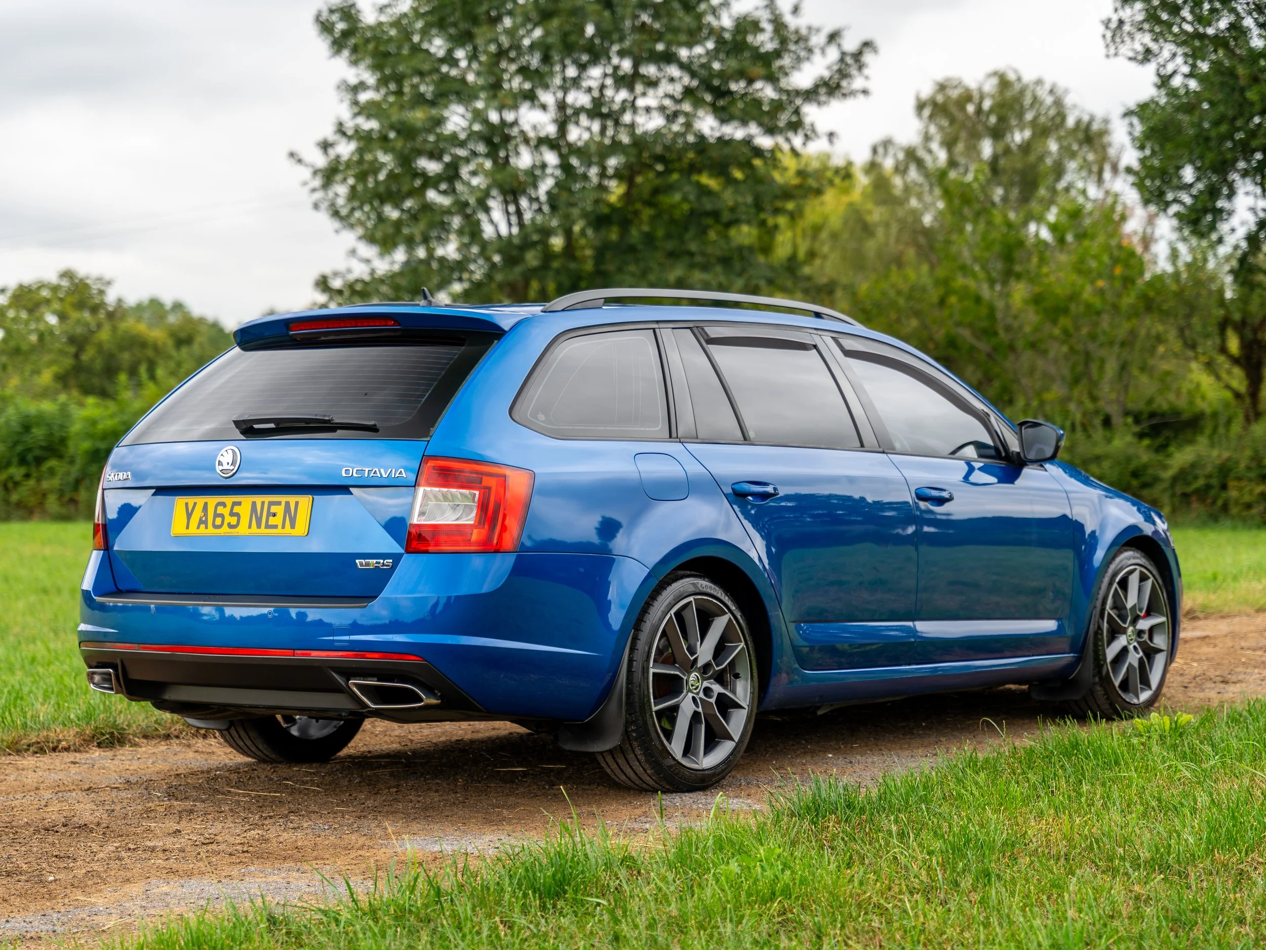 Blue station wagon car parked on dirt path with green grass and trees in the background.