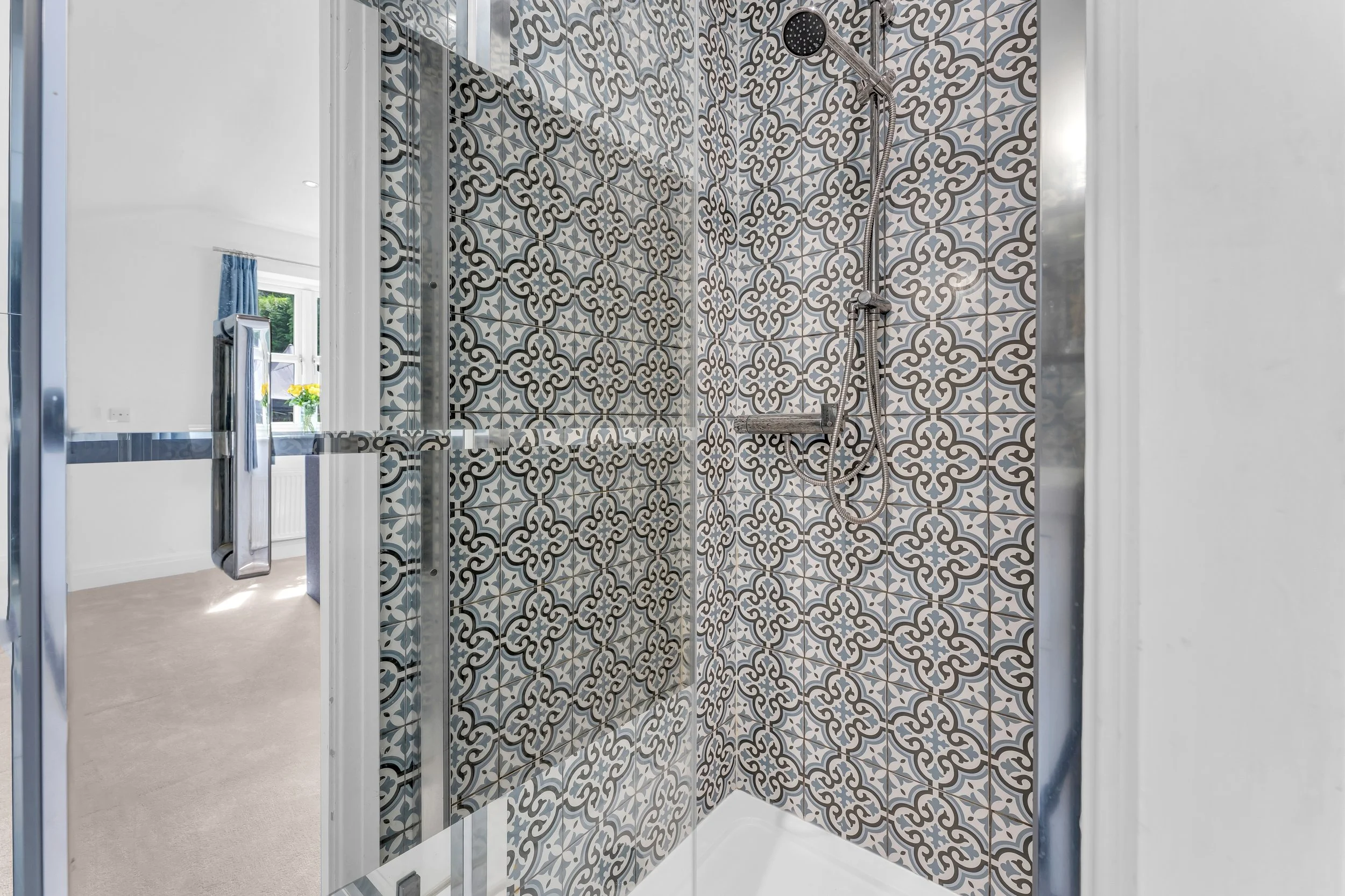 Inside a walk-in shower with decorative patterned tiles on the wall, a handheld showerhead, and a built-in shelf, with the bathroom visible outside through a glass door.