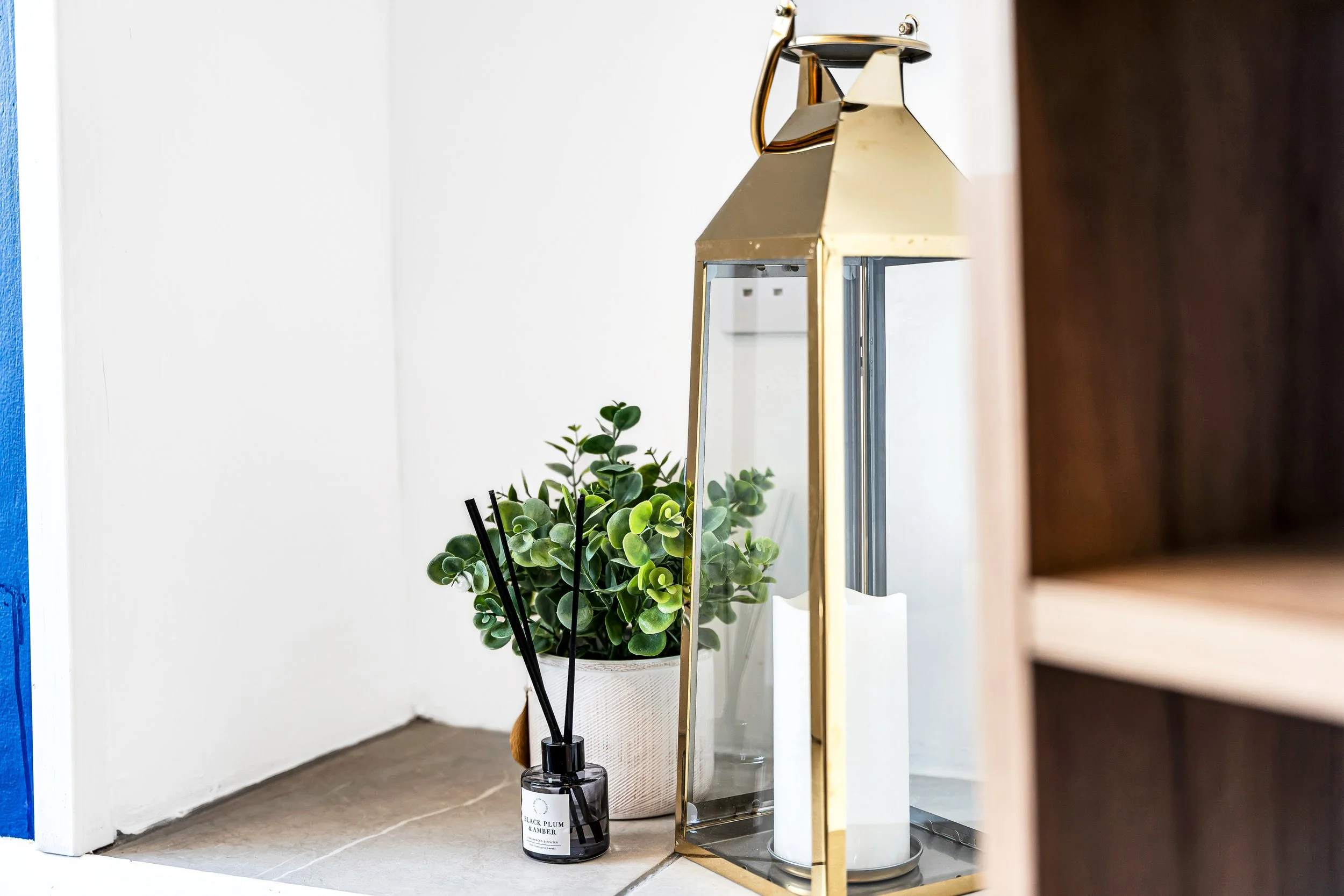 A white pot with green leafy plant, black reed diffuser, and a tall glass lantern with a white candle inside on a tiled floor