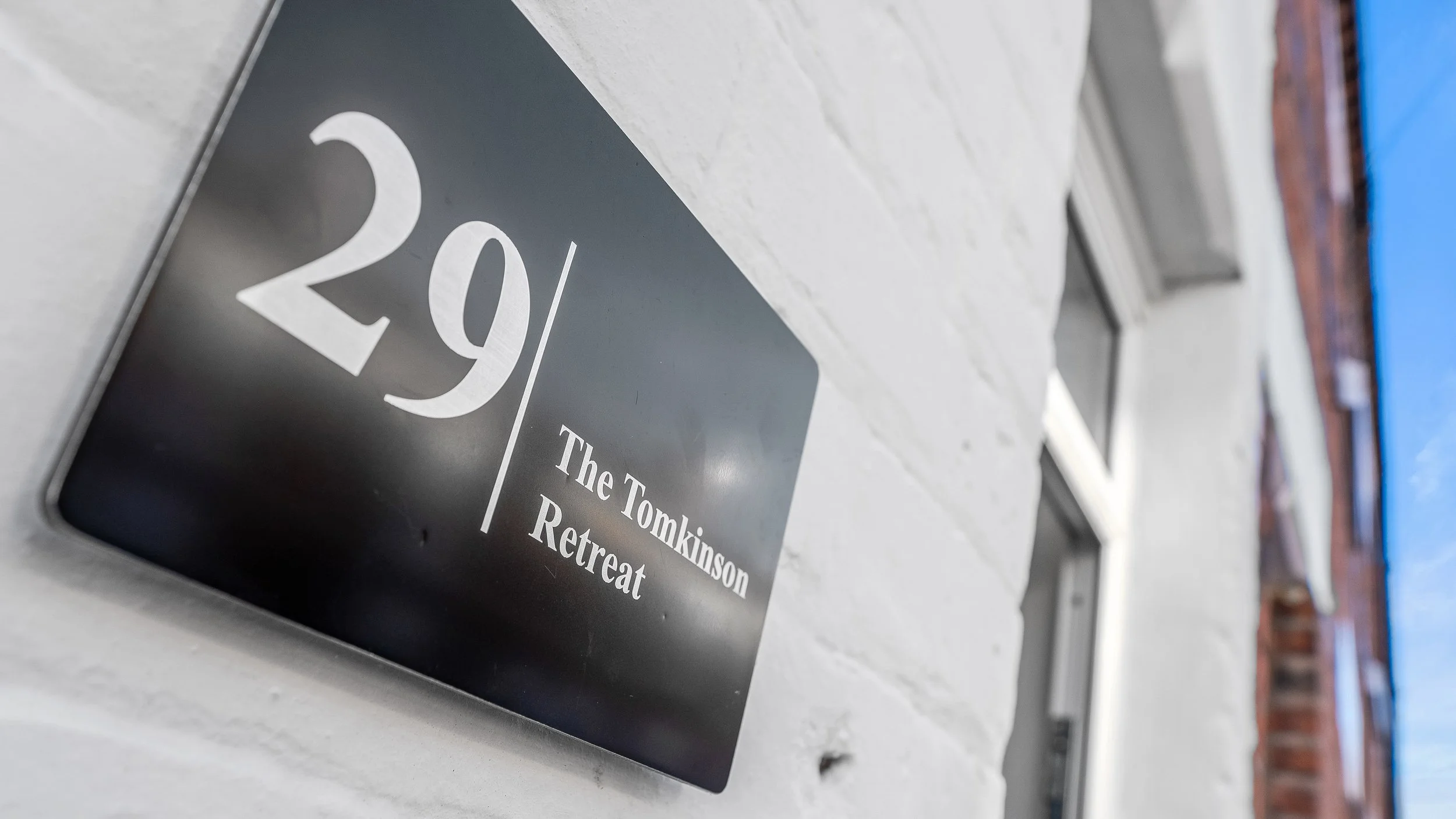 House number 29 / The Tomkinson Retreat sign on a white wall, with windows and a blue sky visible in the background.