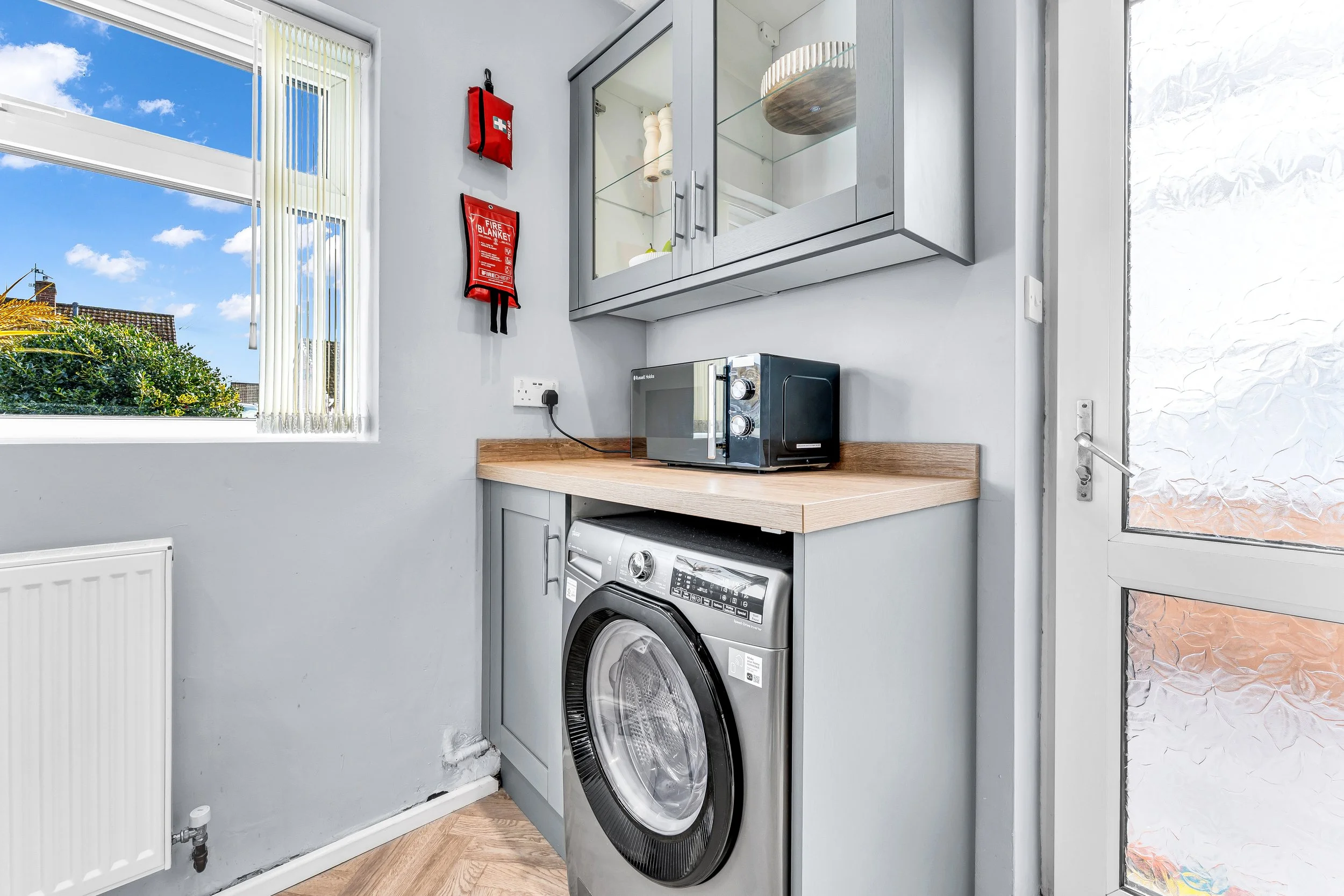 A laundry room with a washing machine under a wooden countertop, a microwave oven, wall-mounted cabinets, a window showing a blue sky with clouds, and a fire blanket and first aid kit on the wall.