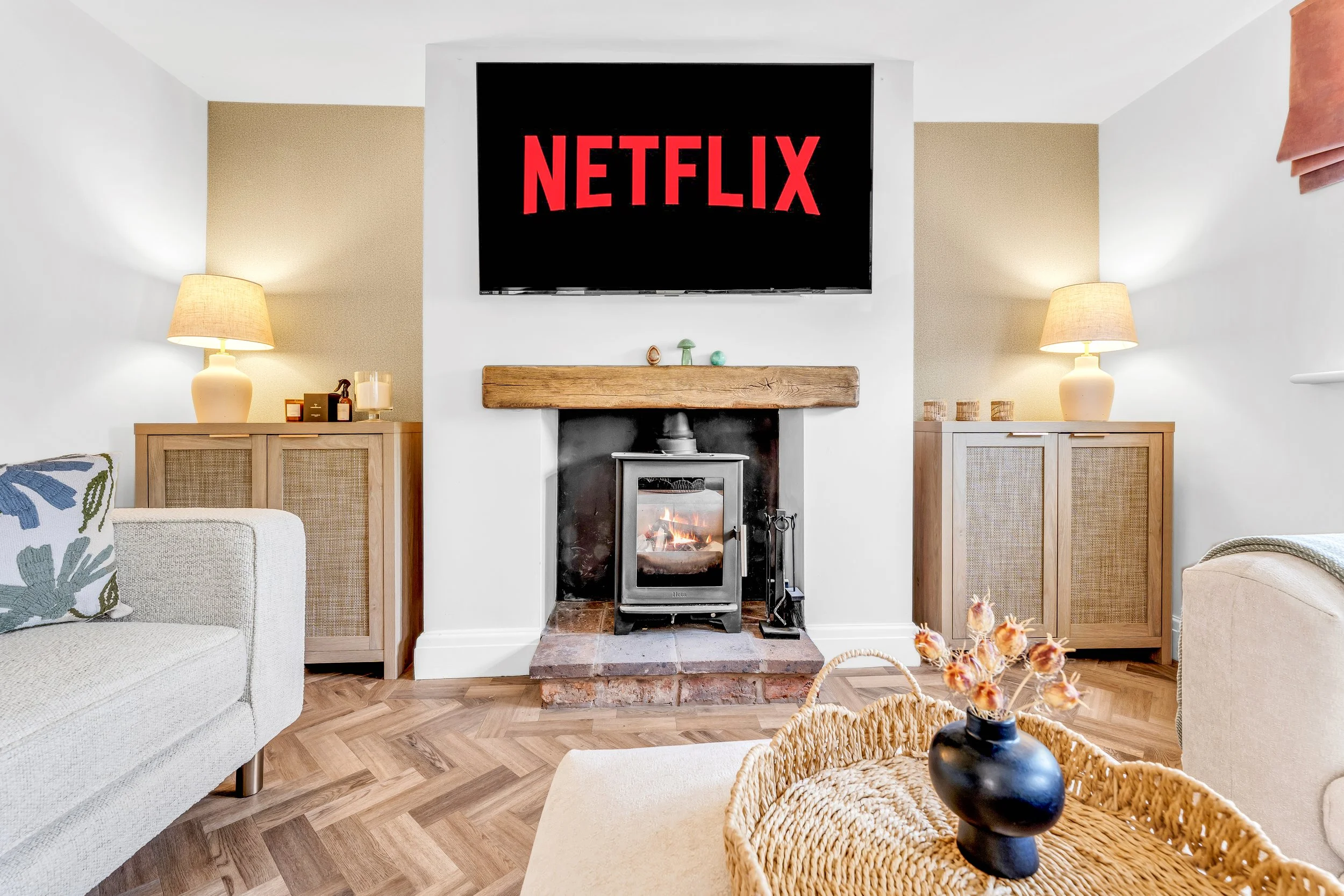 Living room with a fireplace, two side tables with lamps, a large wall-mounted Netflix screen, and furniture including a cream armchair with a patterned pillow and a wicker basket with orange flowers.