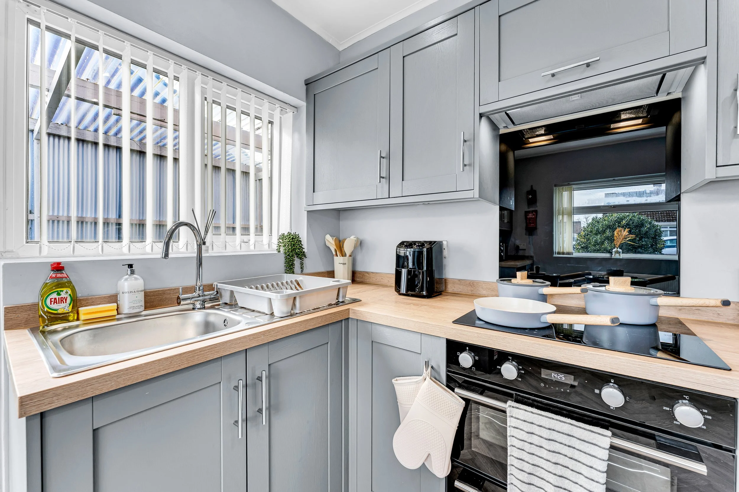 Kitchen with gray cabinets, a window with vertical blinds, and a wooden countertop. Items include dish soap, sponge, dish rack, dishware, and small appliances like a toaster and pots on a stove. There is a window that shows trees outside.