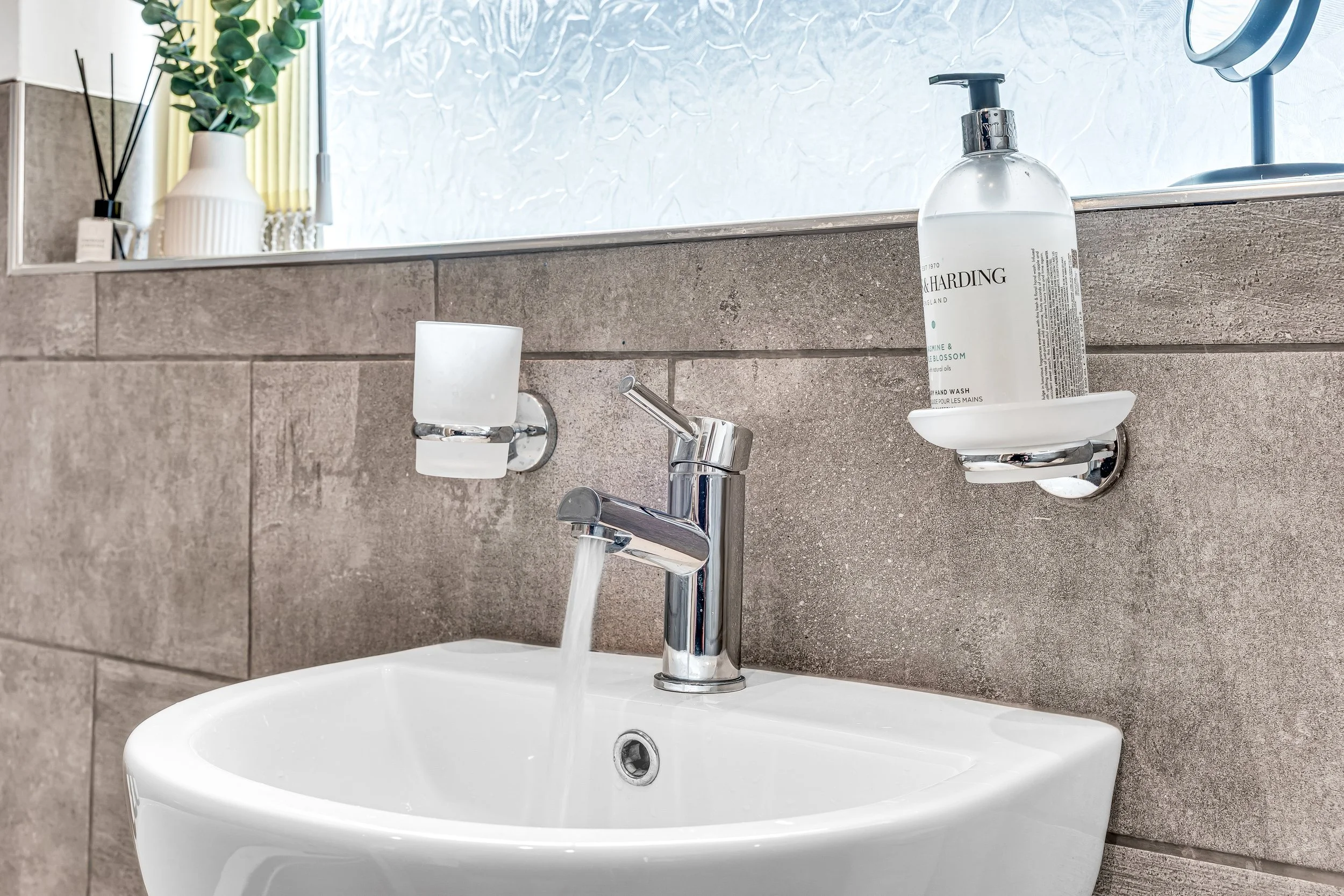 Bathroom sink with a chrome faucet running water, mounted on a beige tiled wall. To the right, a bottle of liquid hand soap on a wall-mounted soap dish, and a square wall-mounted box for tissues or soap. In the background, a window with frosted glass