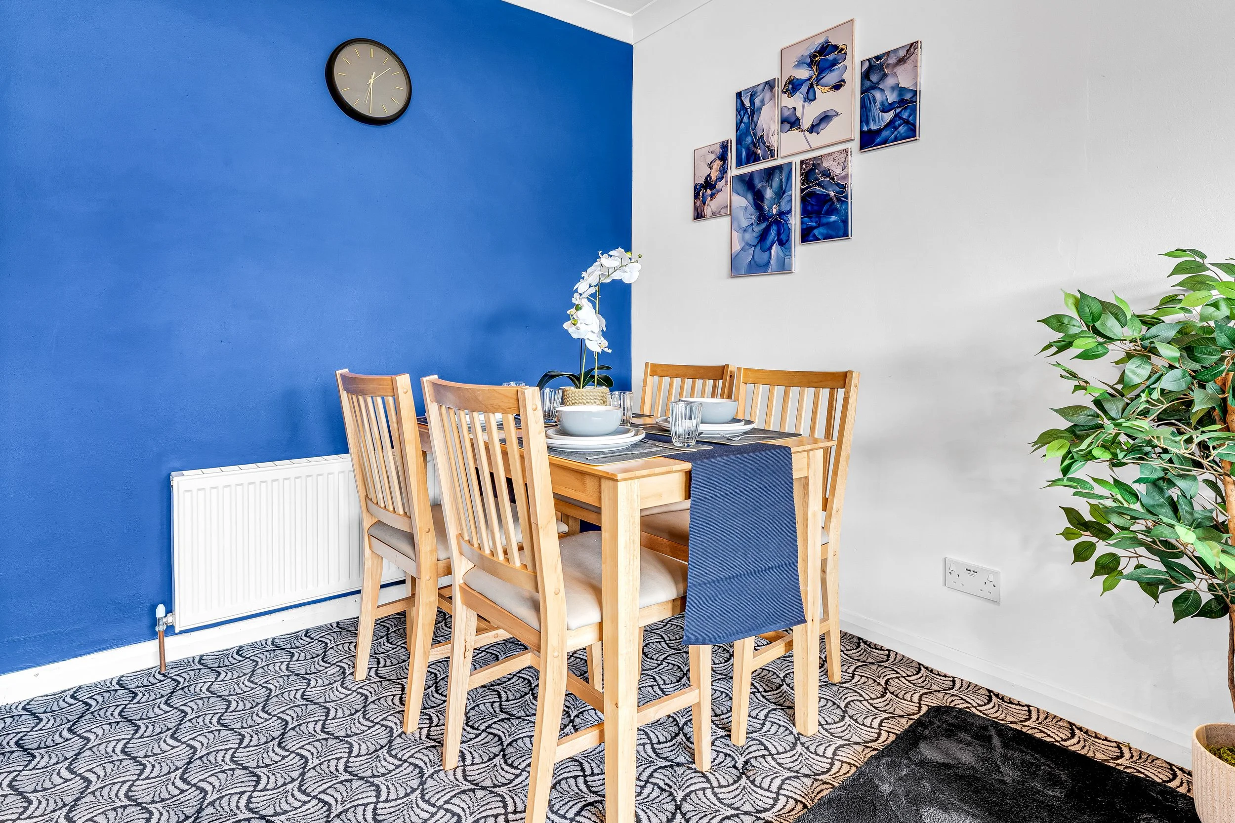 Dining room with a wooden table and six chairs, a blue accent wall with a wall clock, a large green plant, and framed blue abstract artwork.