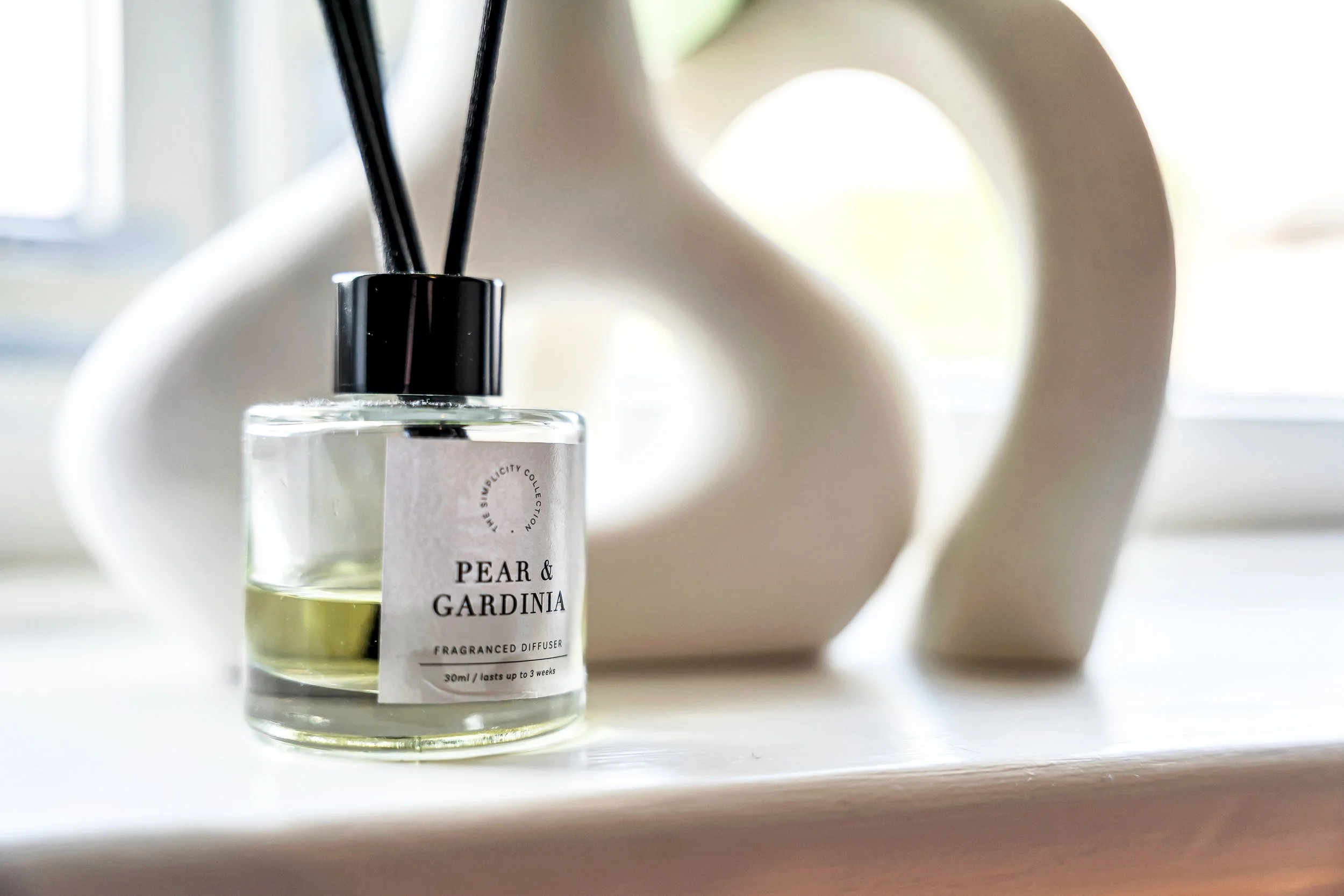 A glass diffuser bottle labeled 'Pear & Gardinia' with a black cap and black reed sticks in it, placed on a white surface next to a white, abstract ceramic sculpture with a hole in it.