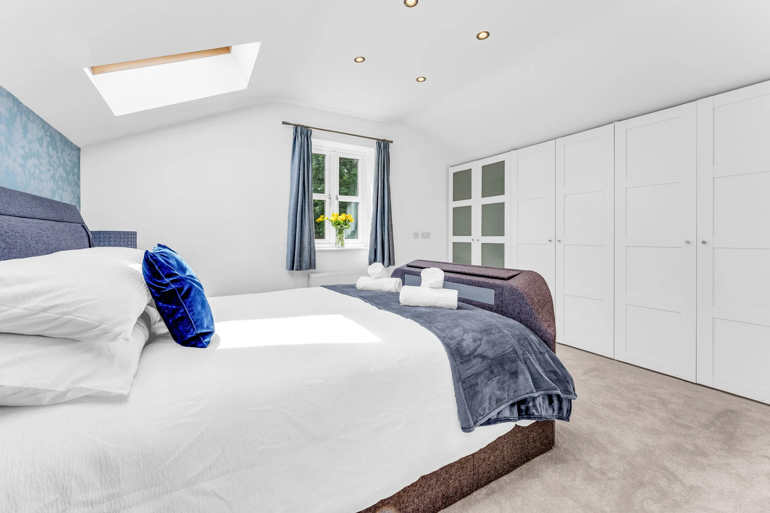 A modern bedroom featuring a large bed with white sheets and blue pillows, a window with blue curtains, a vase of yellow flowers, a skylight, and built-in white wardrobes.