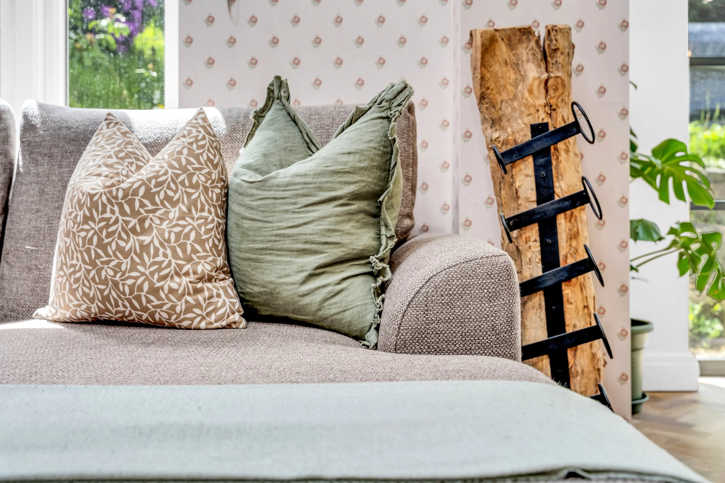 A cozy living room corner with a beige fabric sofa adorned with three pillows, a large window with a view of greenery outside, a rustic wooden coat rack behind the sofa, and a green houseplant to the right.