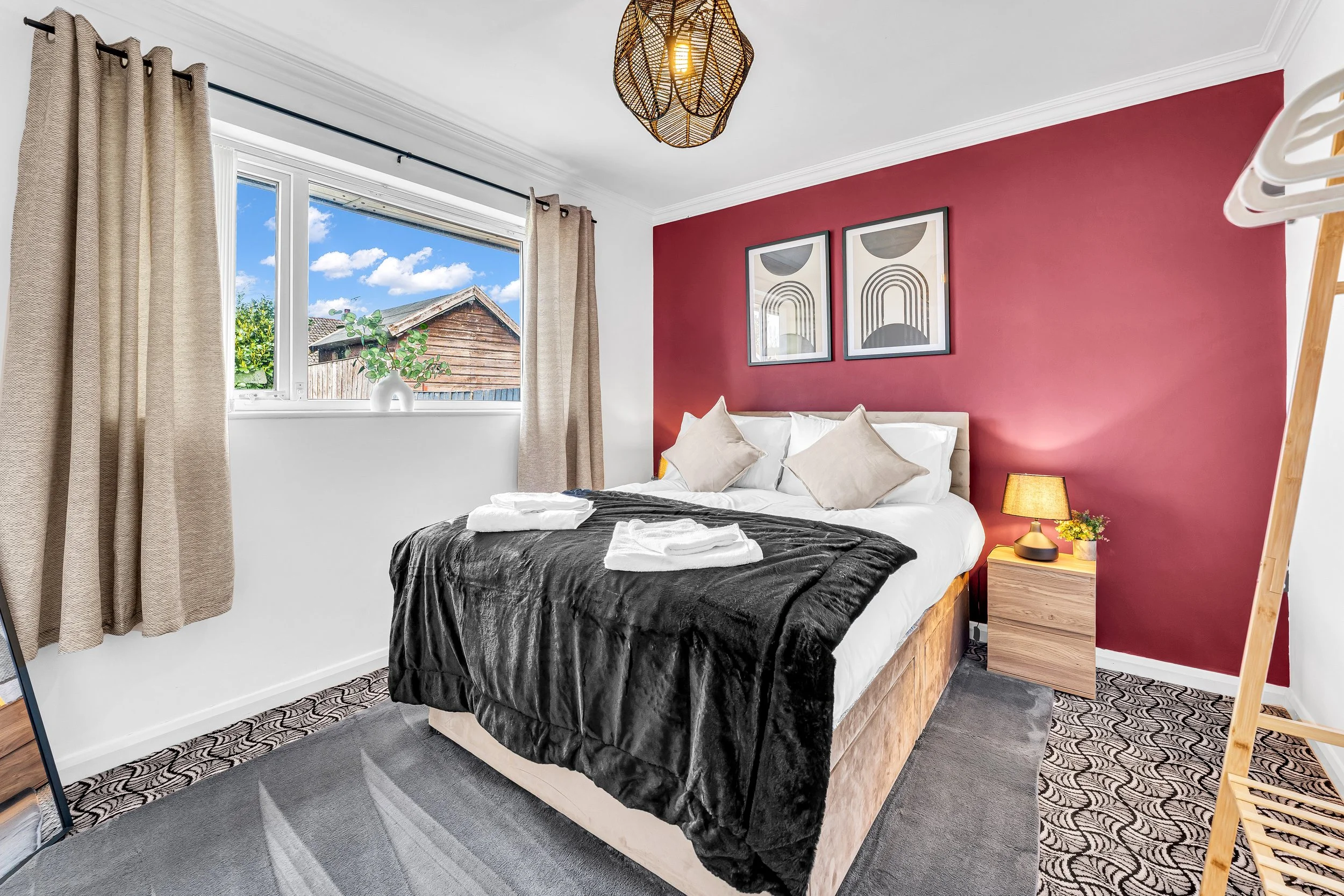 A bedroom with a bed covered in white and black bedding, beige pillows, and two folded towels. There's a pink accent wall with two framed abstract art prints. A nightstand with a lamp and small plant is beside the bed. A window with beige curtains sh