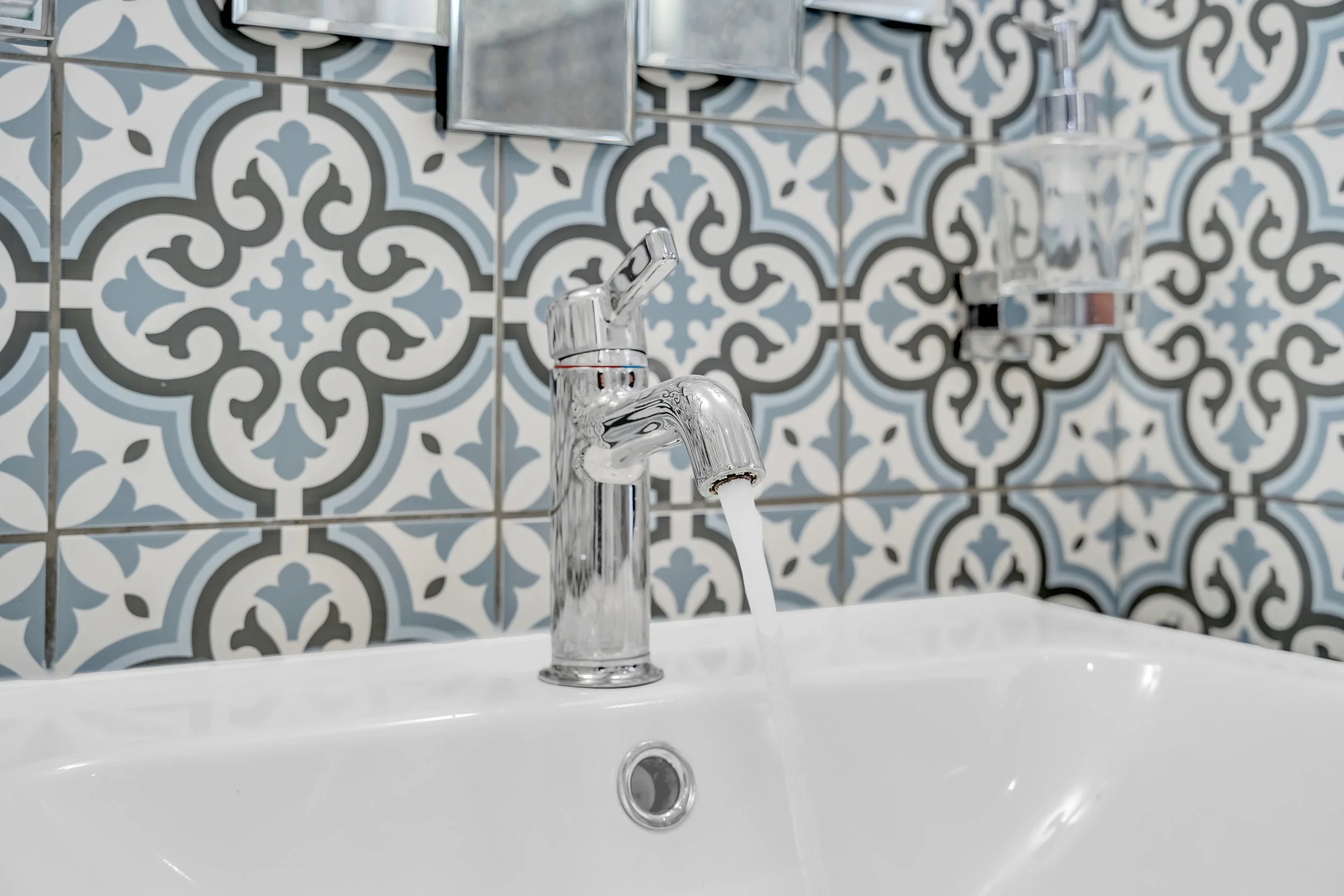 A close-up of a sink with running water, a chrome faucet, and a decorative tiled backsplash with intricate gray, black, and white patterns.