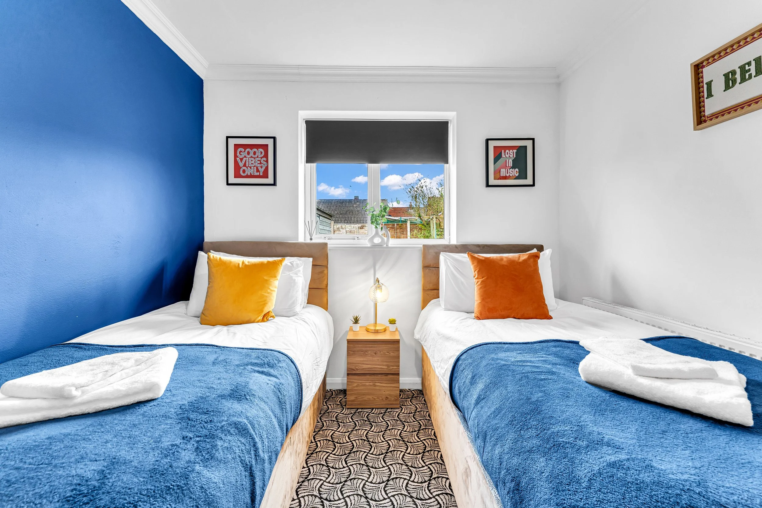 A bedroom with two twin beds, each with white sheets, blue blankets, and a yellow or orange pillow. Between the beds is a wooden nightstand with a small lamp and decorative items. The window above the nightstand shows a view of rooftops and trees out