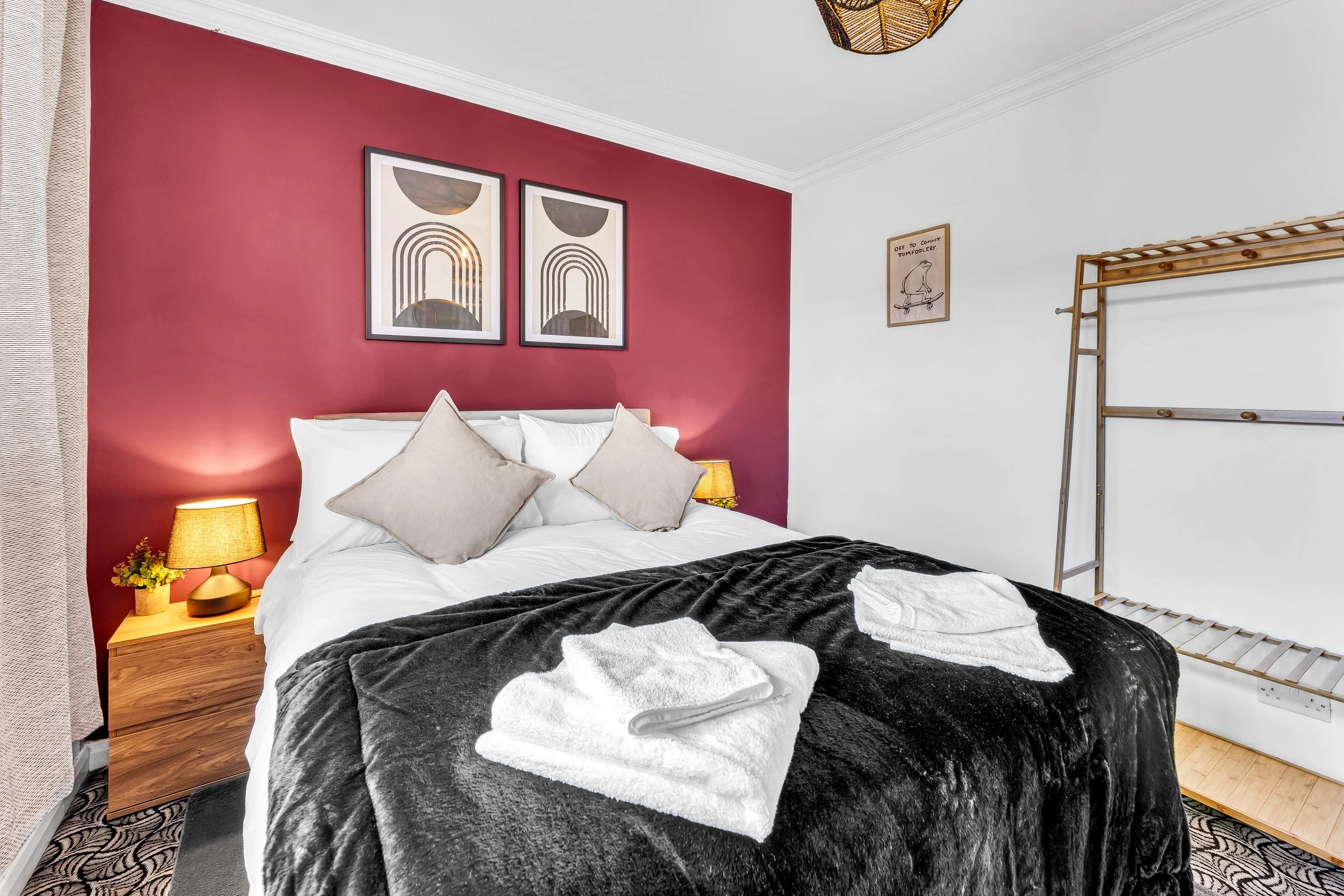 A bedroom with a bed, two lamps, artwork on the walls, and a wooden rack, with a maroon accent wall and white walls.